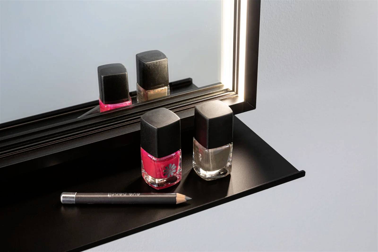 Zwei Nagellackfläschchen, rot und transparent, stehen auf einem schwarzen Regal vor einem Spiegel. Ein schwarzer Stift liegt daneben.