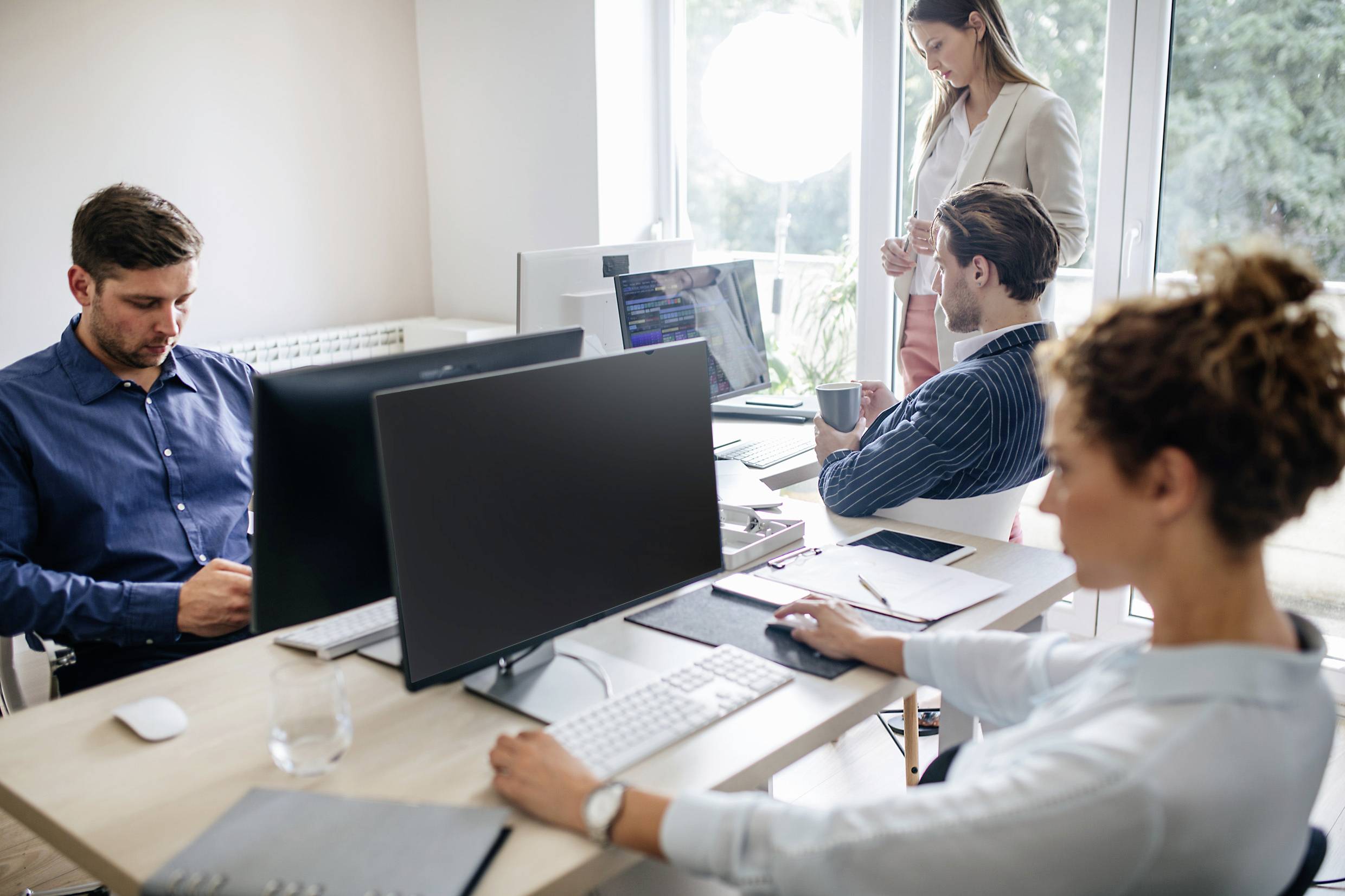 Ein Büro mit vier Personen, die an Computern arbeiten. Eine Person steht und trinkt Kaffee, während drei Personen sitzen und konzentriert arbeiten.