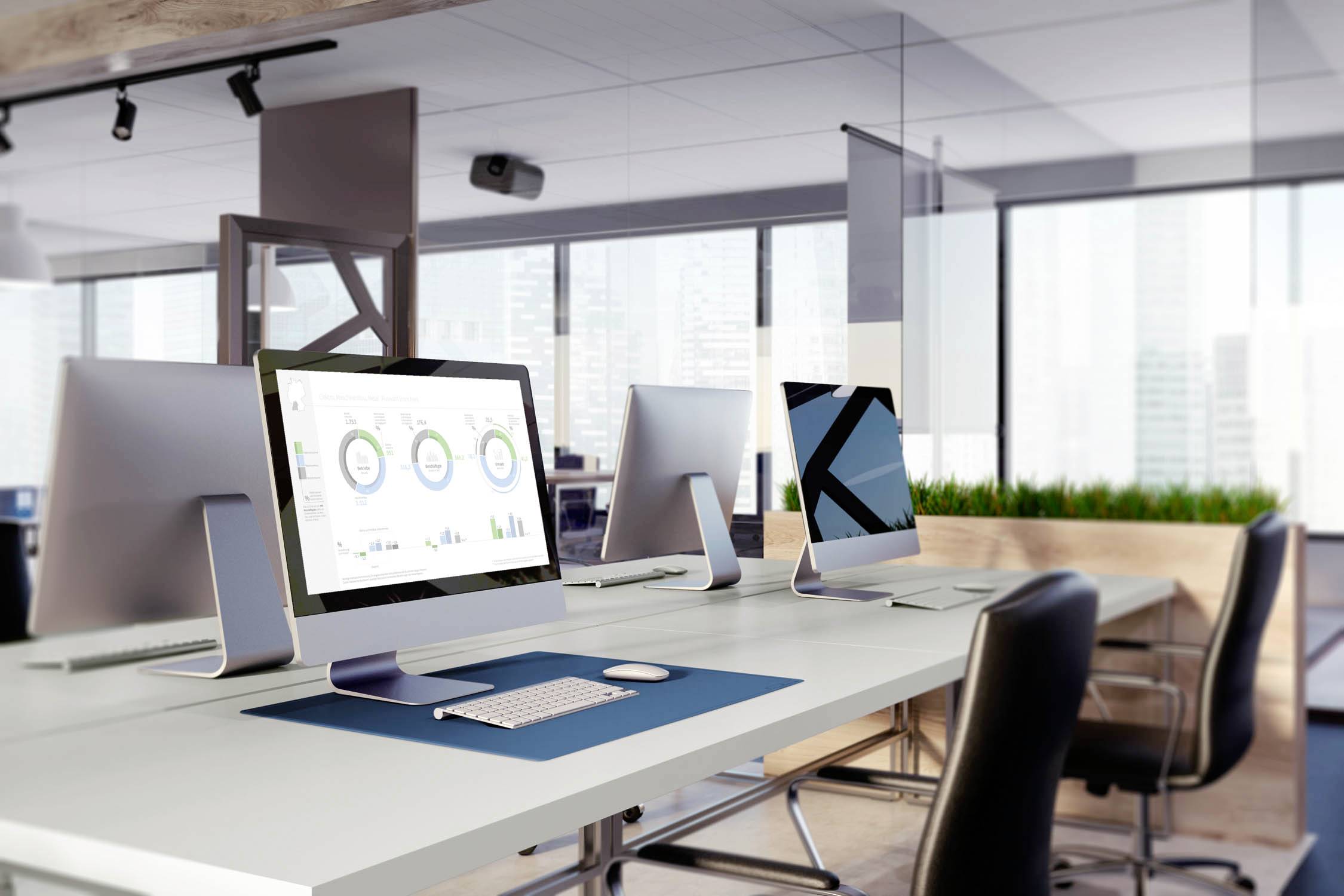 Moderne Büroräume mit mehreren Computern auf Schreibtischen. Auf einem Bildschirm sind Diagramme sichtbar. Große Fenster im Hintergrund.