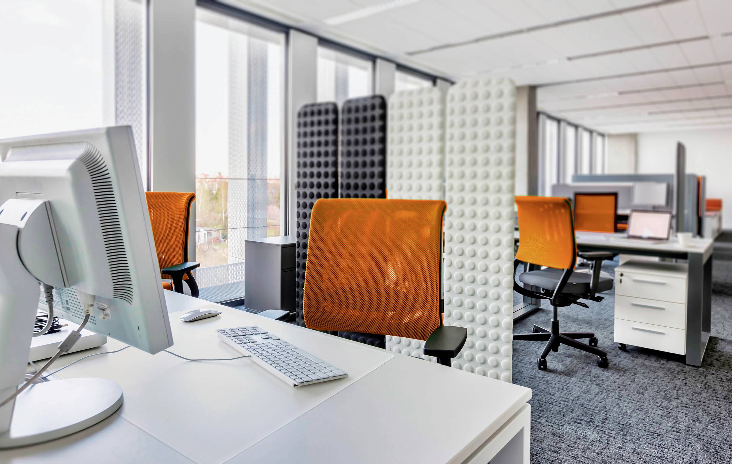 Ein modernes Büro mit weißen Tischen, orangefarbenen Bürostühlen und Trennwänden. Tageslicht fällt durch große Fenster.