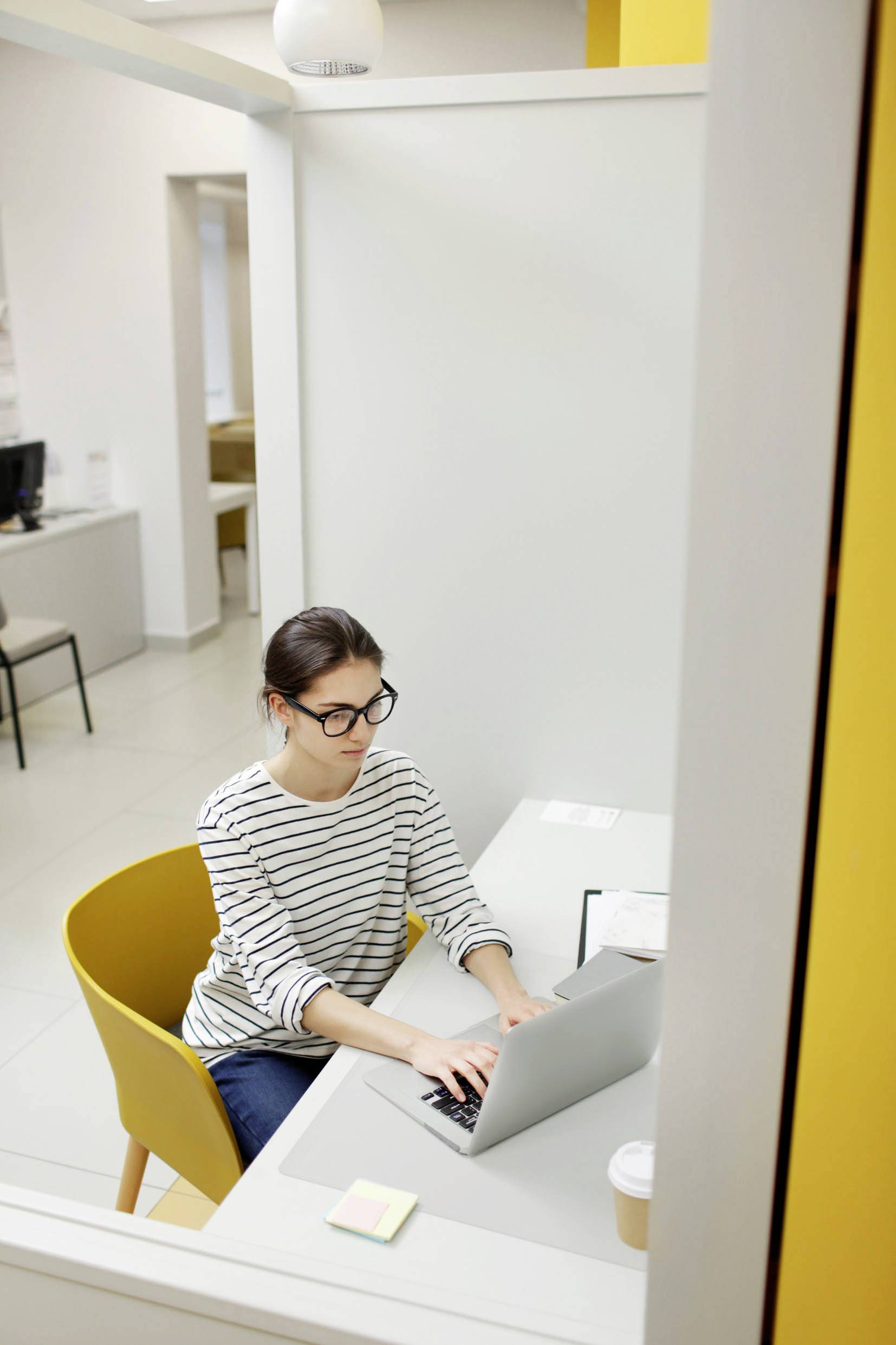 Eine Frau mit Brille arbeitet an einem Laptop in einem Büro, sitzt auf einem gelben Stuhl und hat eine Kaffeetasse auf dem Tisch.