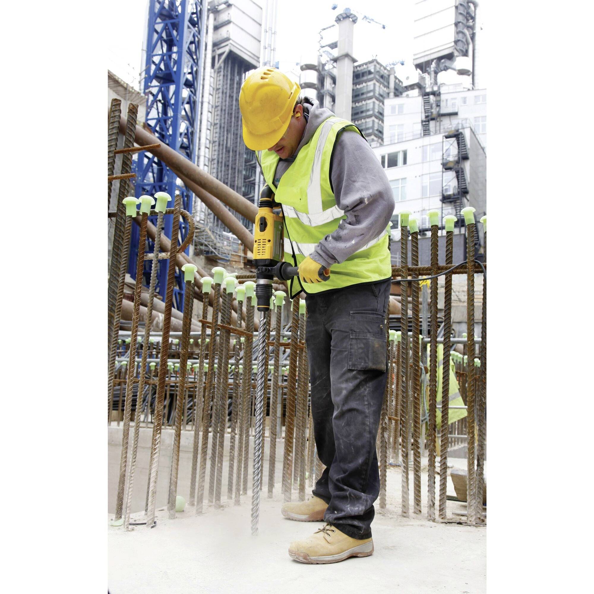 Ein Bauarbeiter bohrt auf einer Baustelle in Beton. Er trägt Schutzkleidung einschließlich Helm und Sicherheitsweste. Im Hintergrund befinden sich Gebäude.