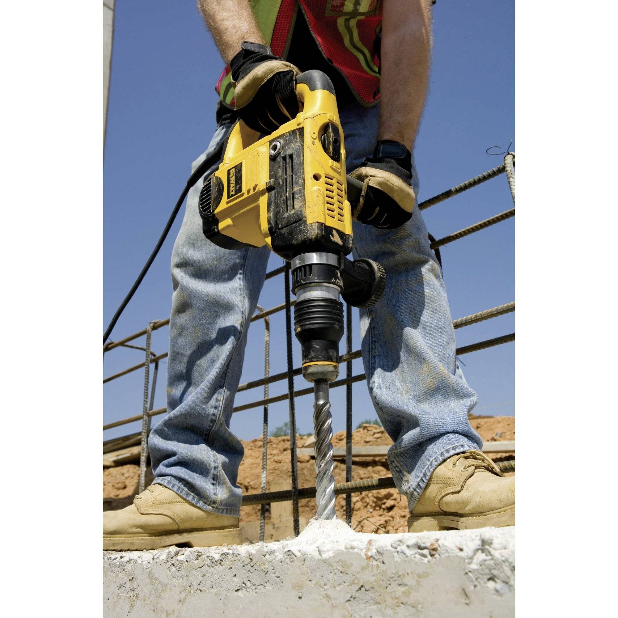 Ein Arbeiter in Jeans und Sicherheitsschuhen bohrt mit einem gelben Elektrowerkzeug in Beton, umgeben von Baustellenmaterialien.