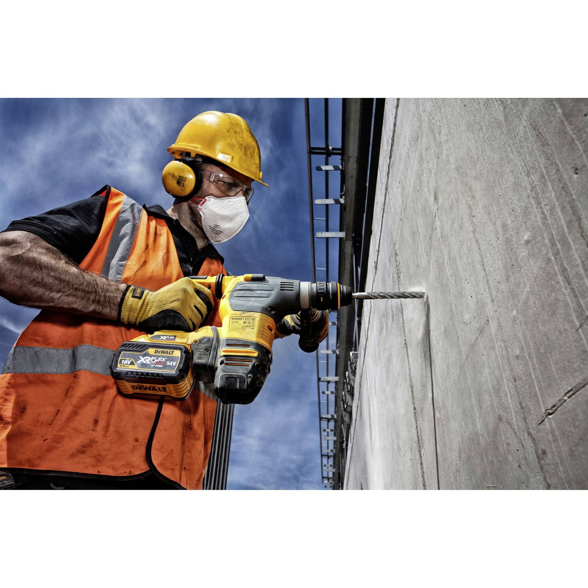 Ein Bauarbeiter in orangener Schutzweste und Helm bohrt mit einer elektrischen Bohrmaschine in eine Betonwand.