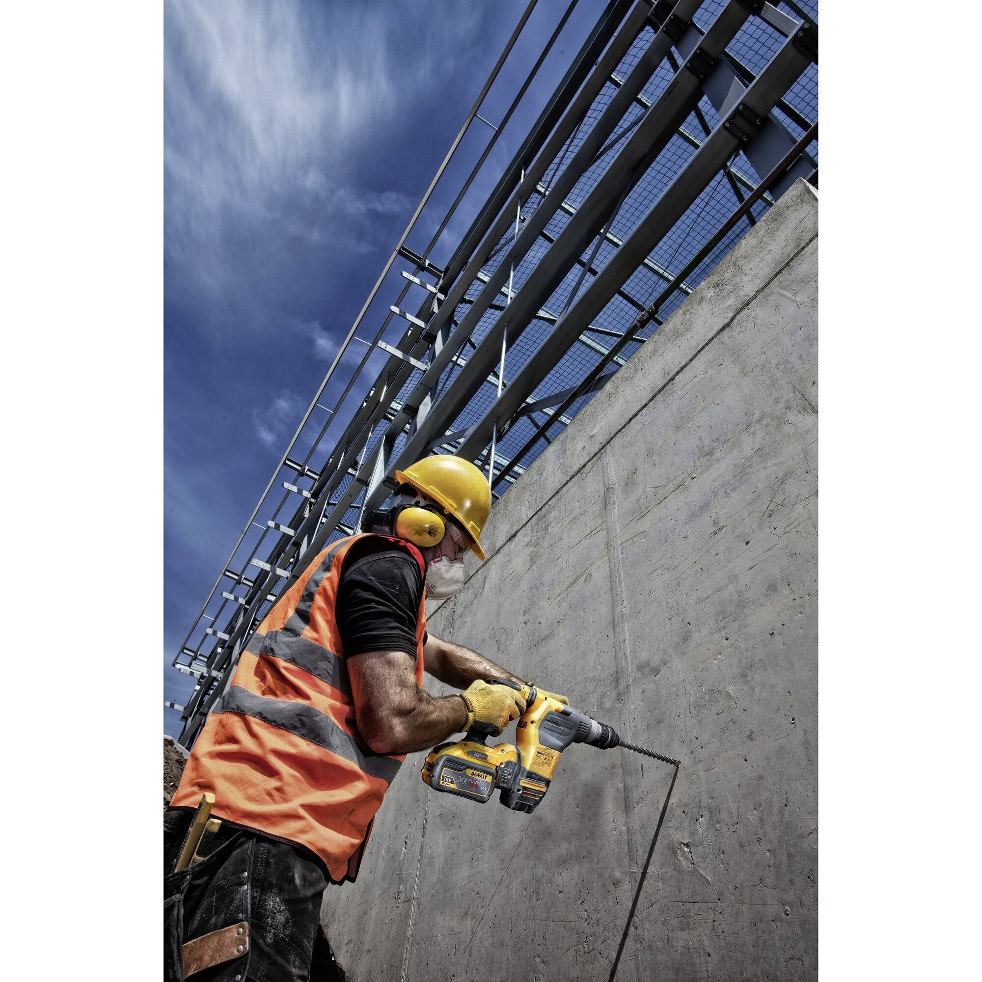 Ein Bauarbeiter trägt eine orangefarbene Weste und einen gelben Helm und bohrt mit einer Bohrmaschine in eine Betonwand unter blauem Himmel.