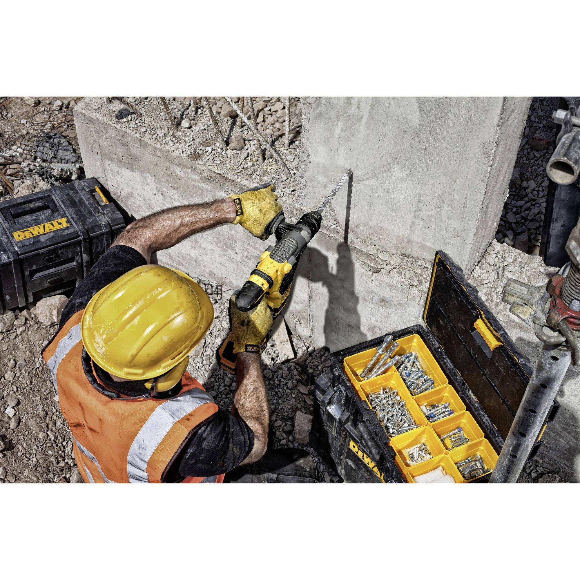 Ein Bauarbeiter mit gelbem Helm und Sicherheitsweste bohrt mit einer gelben Bohrmaschine in eine Betonwand. Werkzeugkasten steht daneben.
