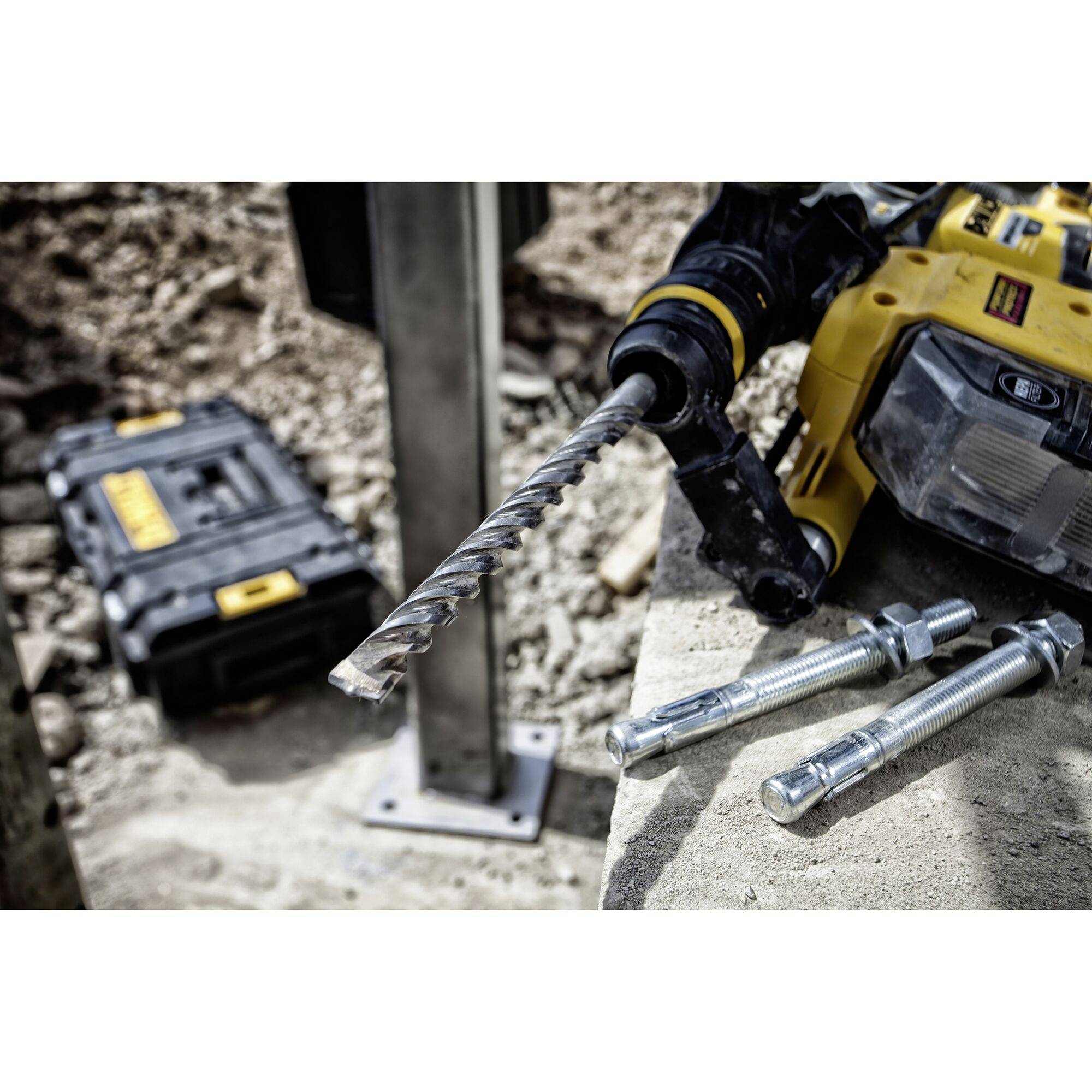 Ein gelber Bohrhammer mit montiertem Bohrer liegt auf einer Betonfläche neben Dübeln. Im Hintergrund ist Werkzeug sichtbar.