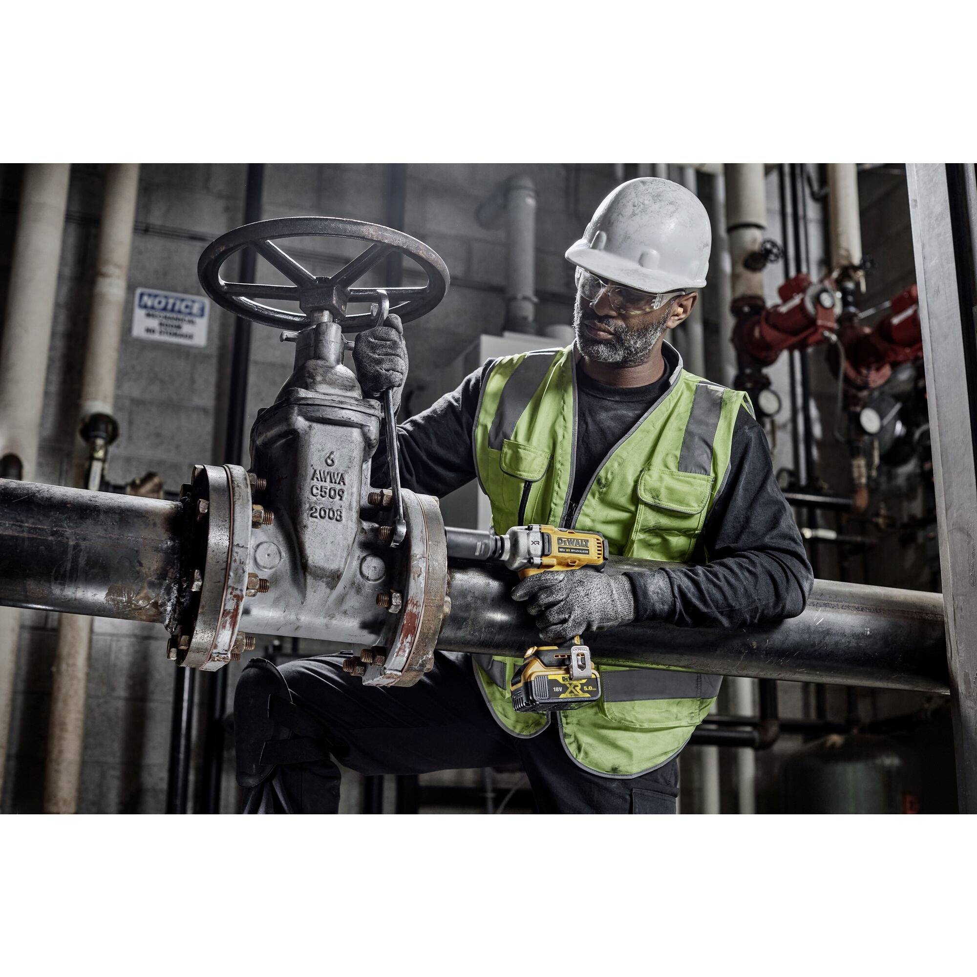 Ein Arbeiter mit Schutzhelm und Warnweste bedient ein großes Rohrventil in einer Industrieanlage, umgeben von Maschinen und Rohren.