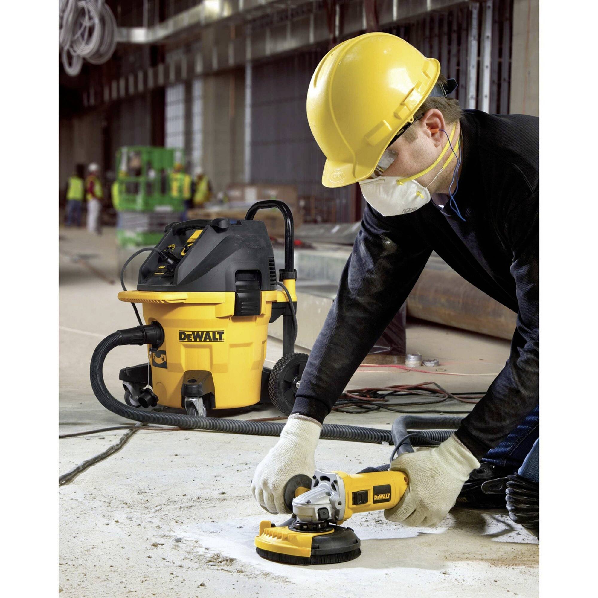 Ein Arbeiter trägt Schutzkleidung und benutzt ein Elektrowerkzeug zum Schleifen einer Betonoberfläche, im Hintergrund ein Industriesauger.