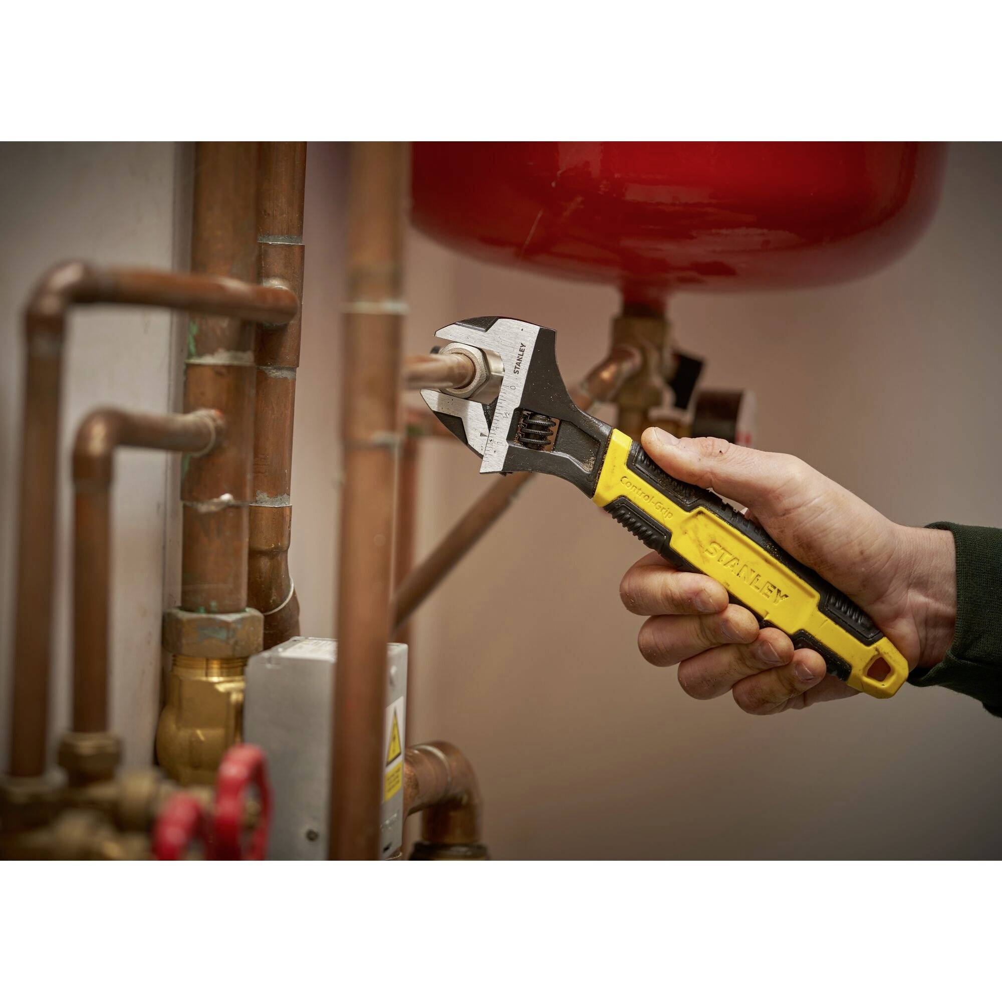 Eine Person zieht eine verstellbare Zange an einem Metallrohr an, umgeben von Kupferleitungen. Technische Arbeiten werden ausgeführt.