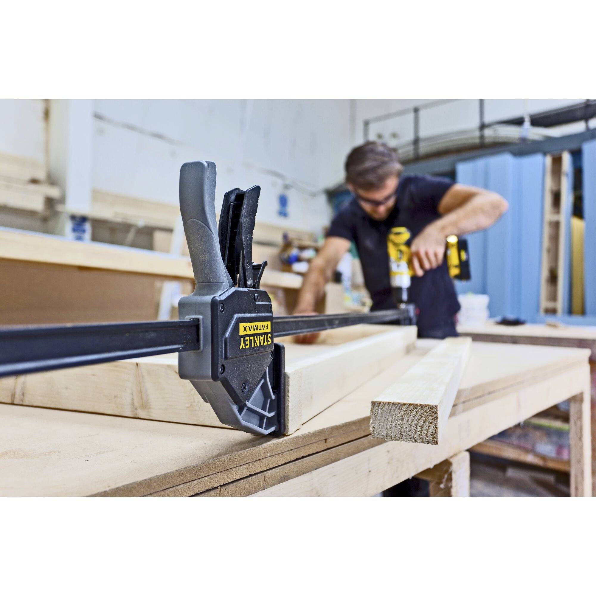 Ein Mann arbeitet in einer Werkstatt, nutzt eine Klammer, um Holzstücke auf einem Tisch zu fixieren. Er trägt Schutzbrille und Werkzeugtasche.