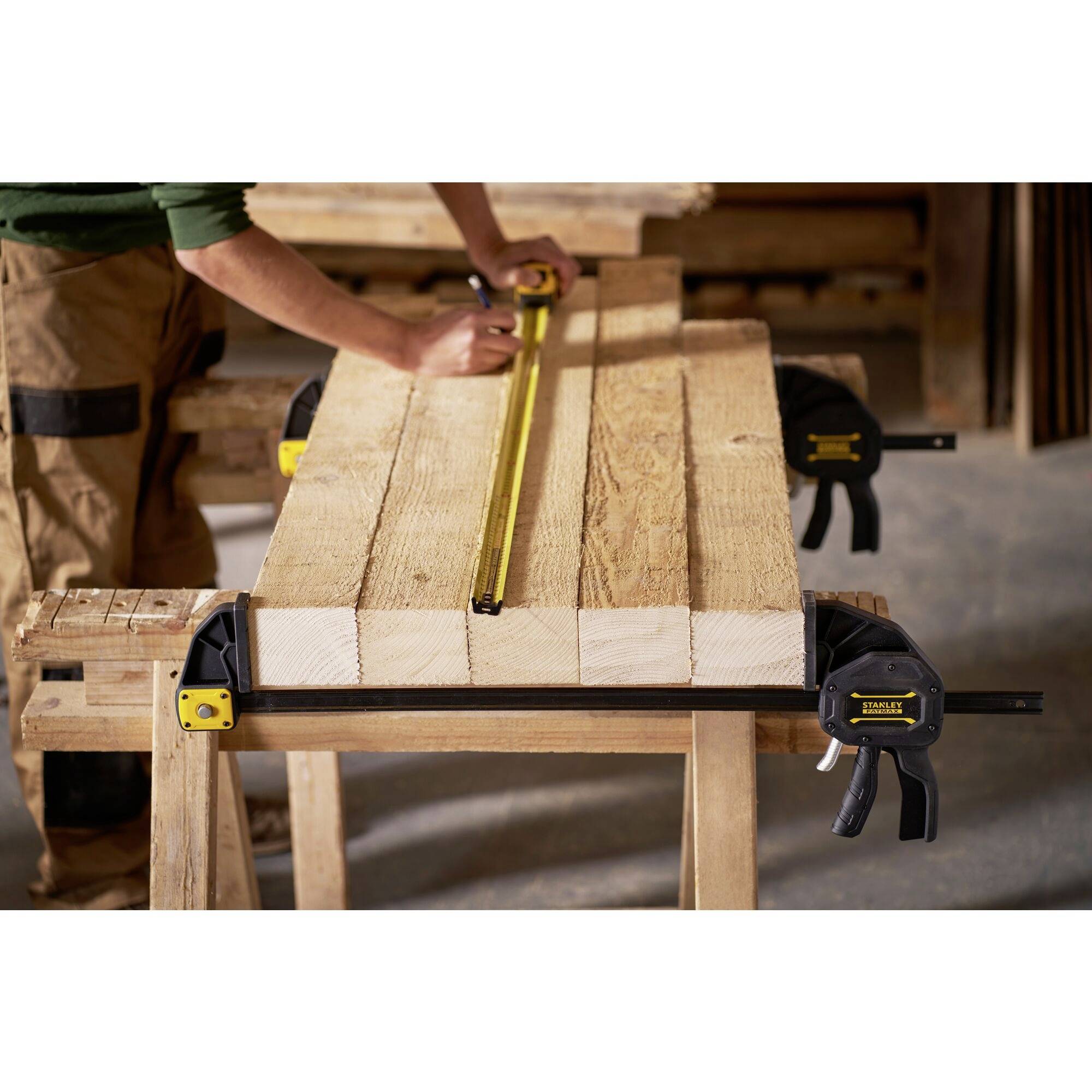 Eine Person misst mit einem Maßband die Länge von Holzbrettern, die auf einer Werkbank mit Klemmen befestigt sind. Arbeitsplatz in einer Werkstatt.