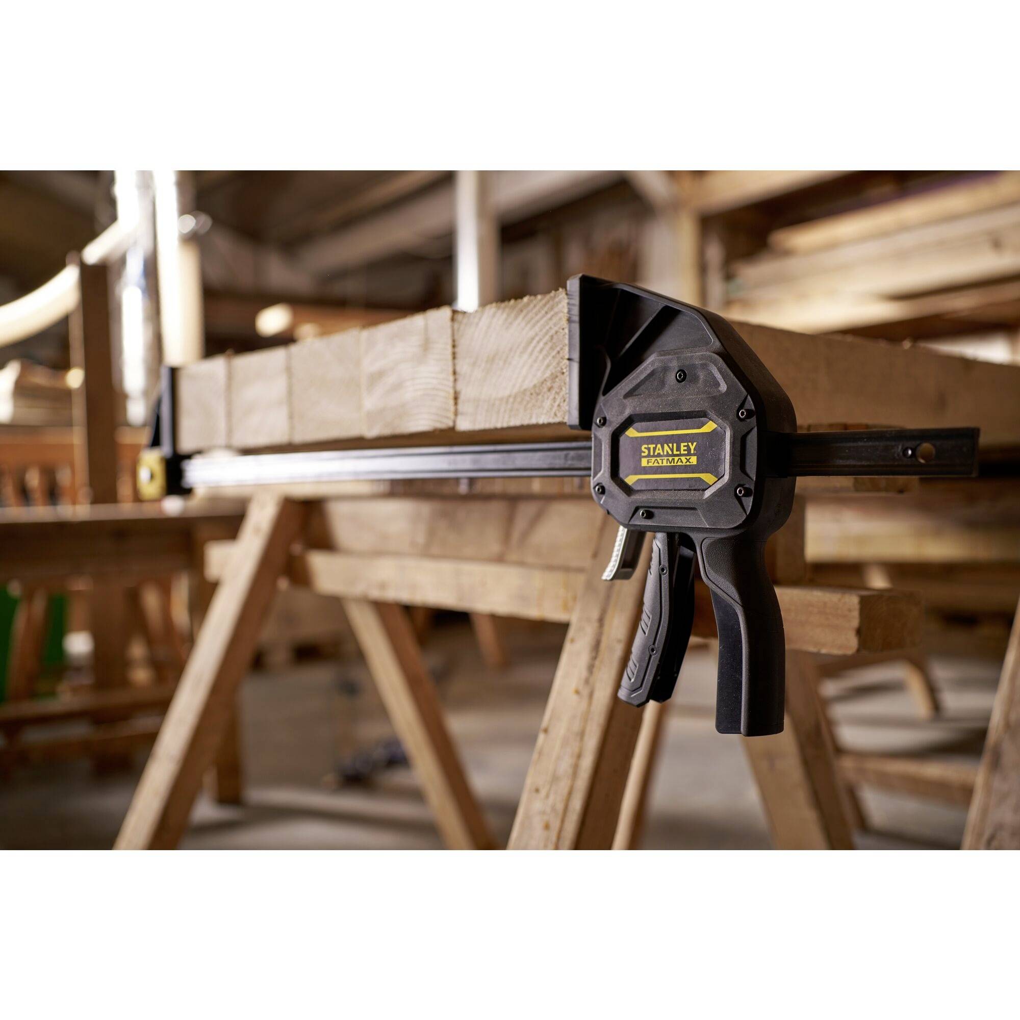 Eine Werkstatt mit einem Holzstück auf einer Werkbank, fixiert durch eine Stanley Zwinge. Hintergrund zeigt unscharfe Holzbearbeitungswerkzeuge.