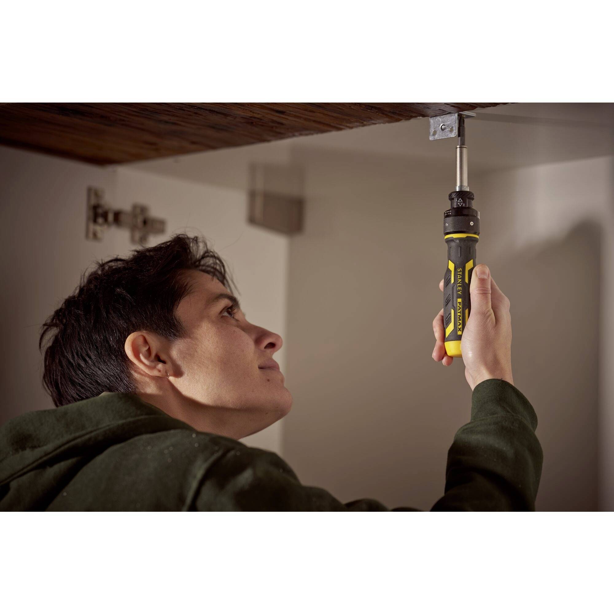 Mann verwendet einen elektrischen Schraubendreher, um eine Schraube in ein Metallstück an der Decke zu befestigen. Er trägt einen Pullover.