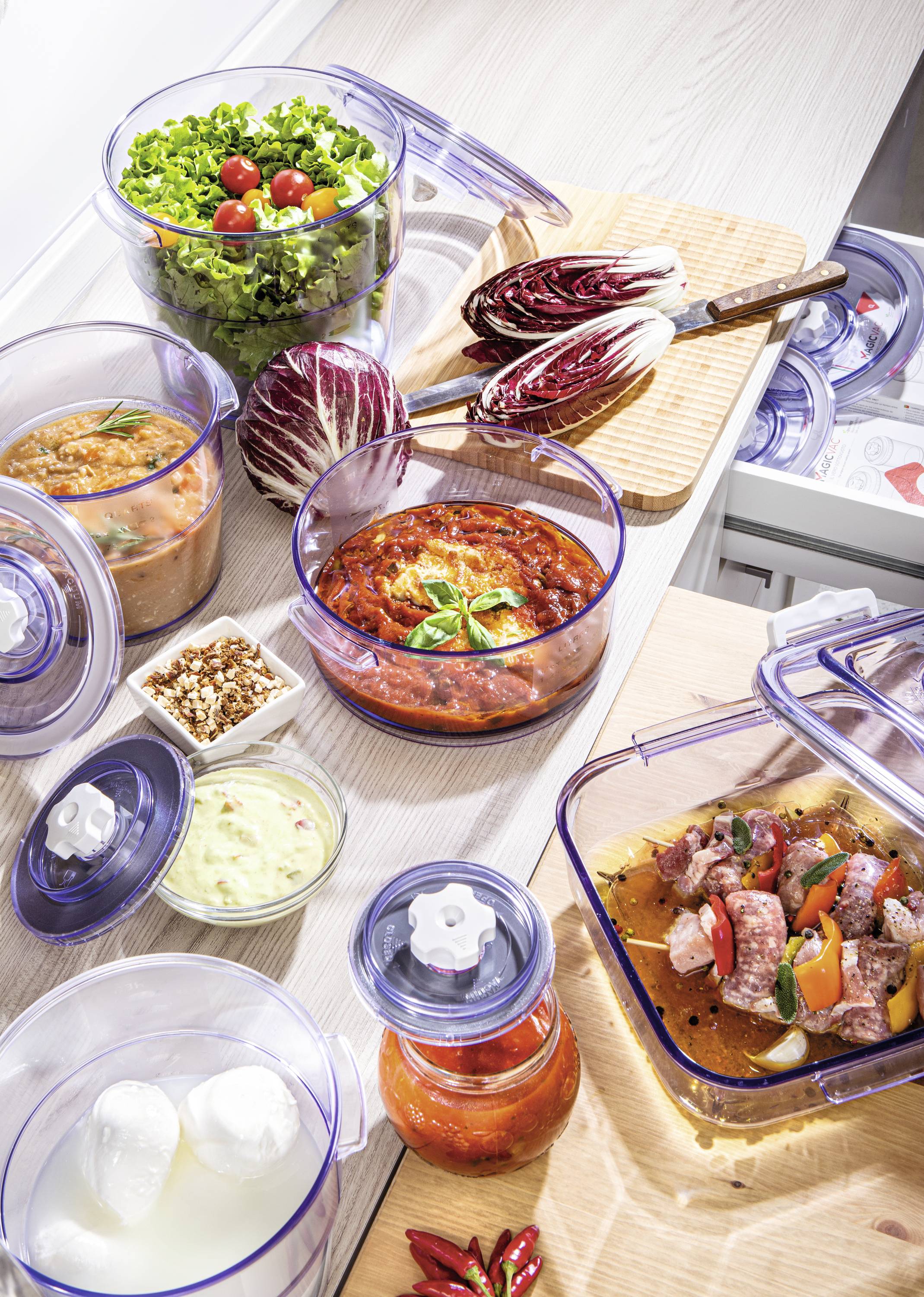 Frische Zutaten und zubereitete Speisen in transparenten Behältern, einschließlich Salat, Tomatensauce und Fleisch, auf einer Küchenarbeitsplatte.