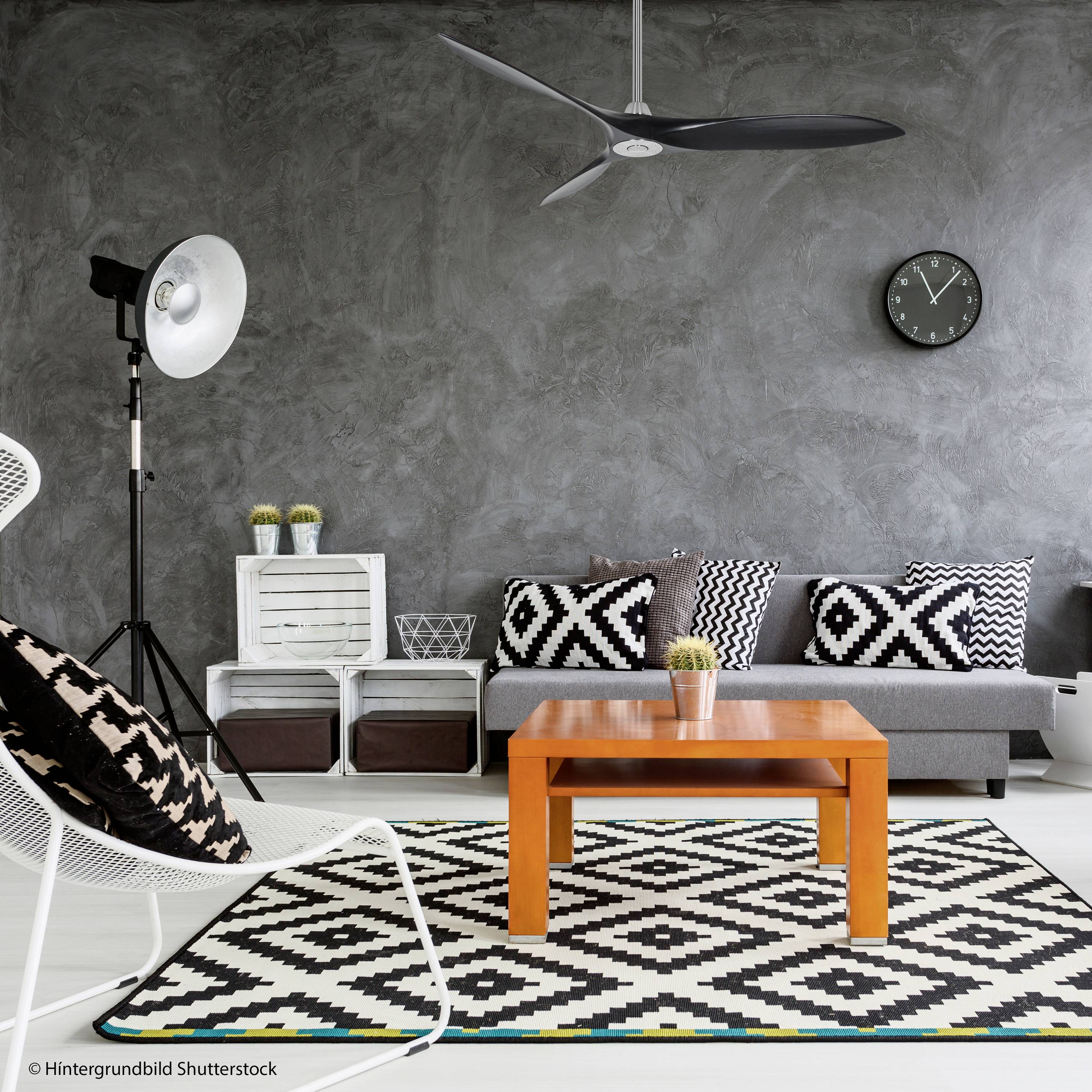 Modern eingerichtetes Wohnzimmer mit grauem Sofa, gemusterten Kissen, orangenem Tisch und schwarz-weißem Teppich. An der Wand eine Uhr.