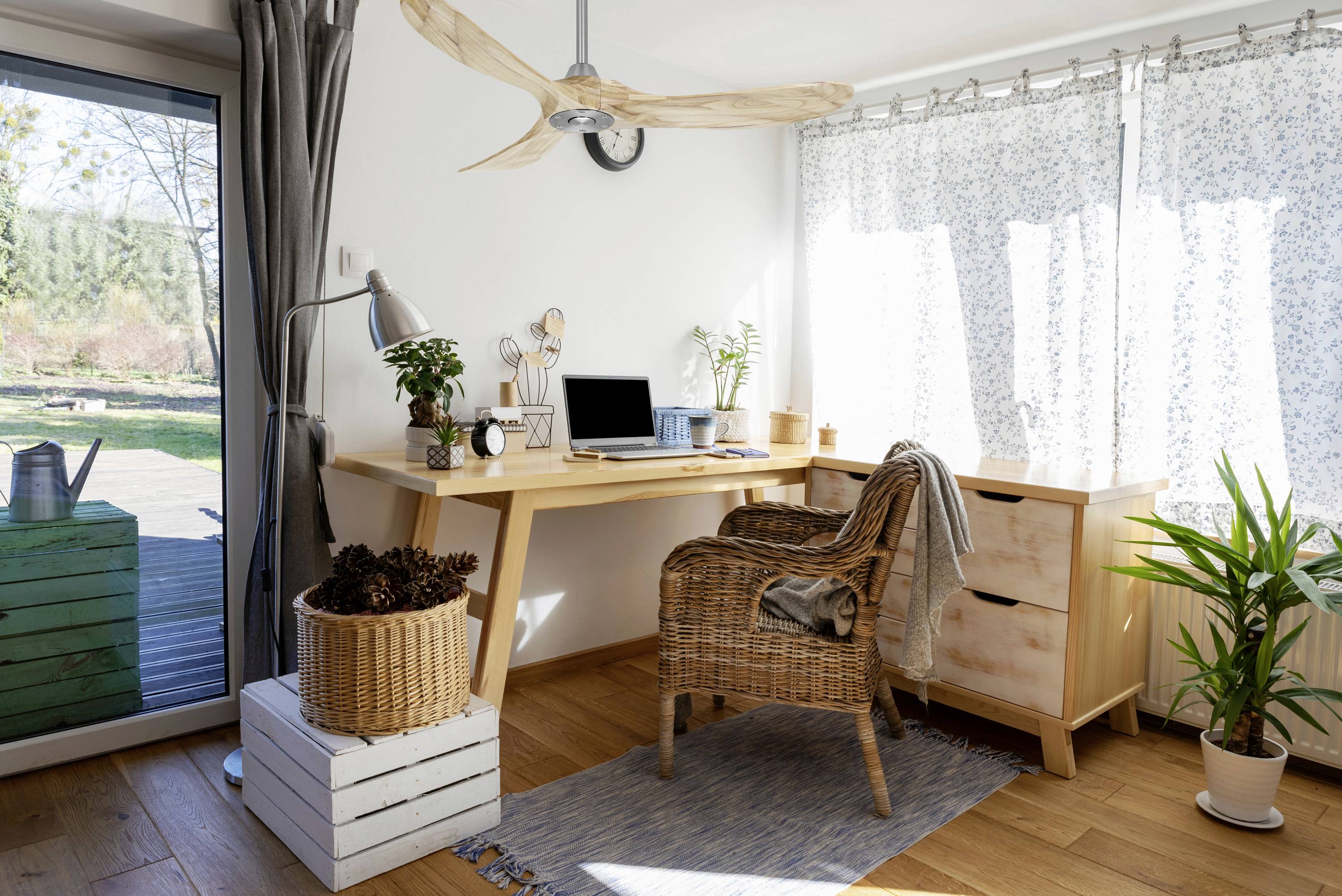 Helles Homeoffice mit Holztisch, Laptop, Korbsessel und Pflanzen am Fenster. Ländliche Außenansicht durch die Glastür.