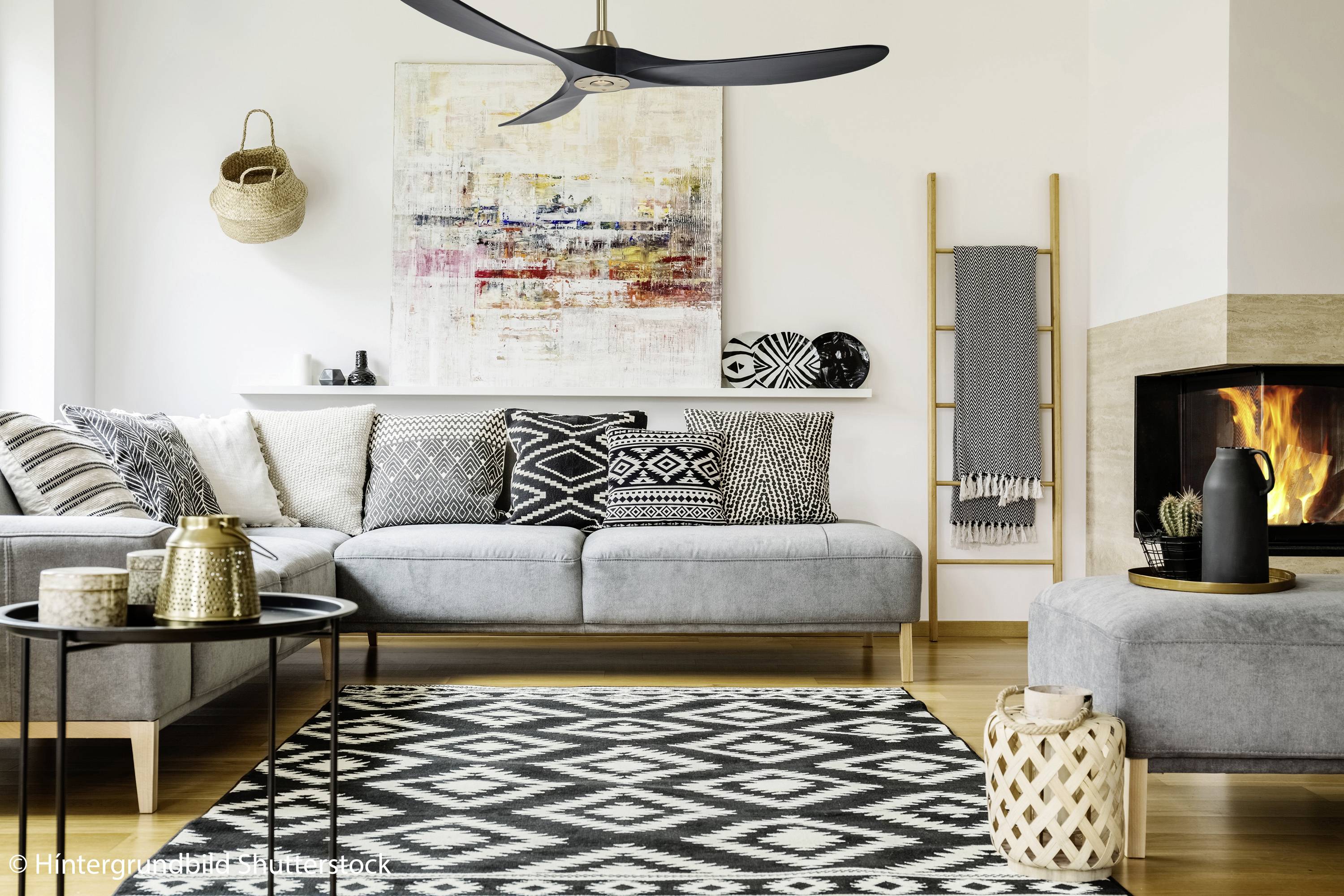 Gemütliches Wohnzimmer mit grauem Sofa, gemusterten Kissen, Teppich, Kaminfeuer, Leiterregal und modernem Deckenventilator.