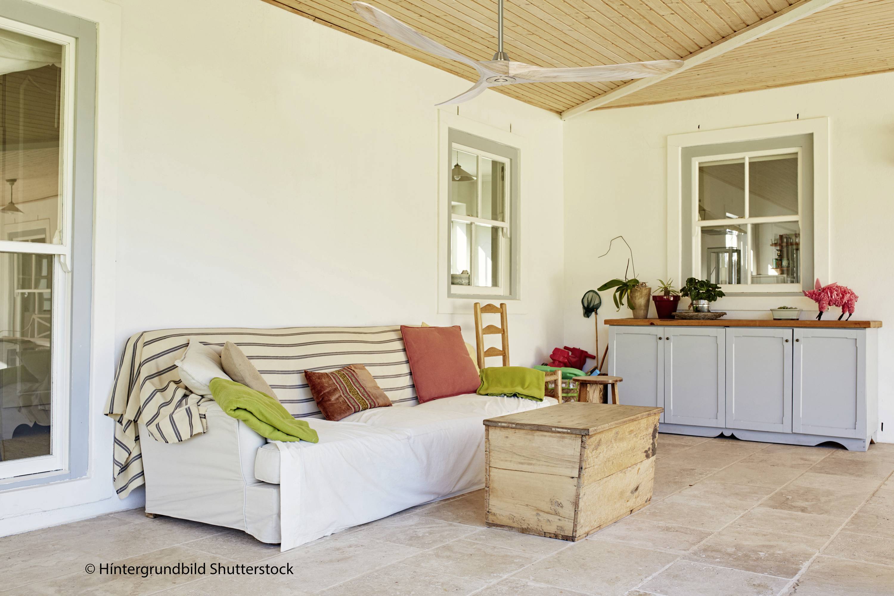 Ein gemütlicher Innenhof mit einem weißen Sofa, farbigen Kissen, einem Holztisch und Pflanzen auf einem Sideboard vor einer weißen Wand.