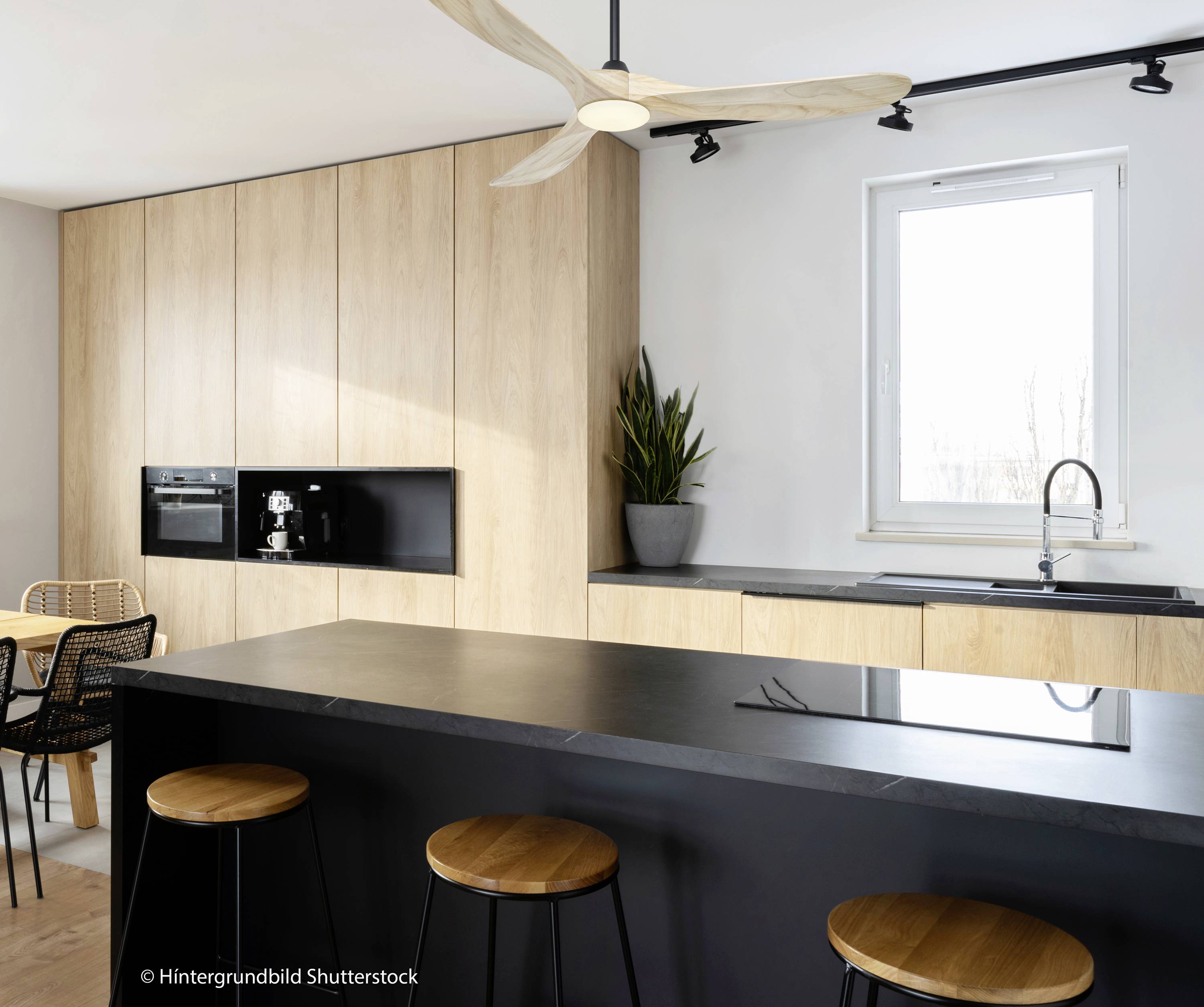 Eine moderne Küche mit Holzelementen, schwarzer Arbeitsplatte, Hockern und einem Fenster. Eine Pflanze steht neben der Spüle.