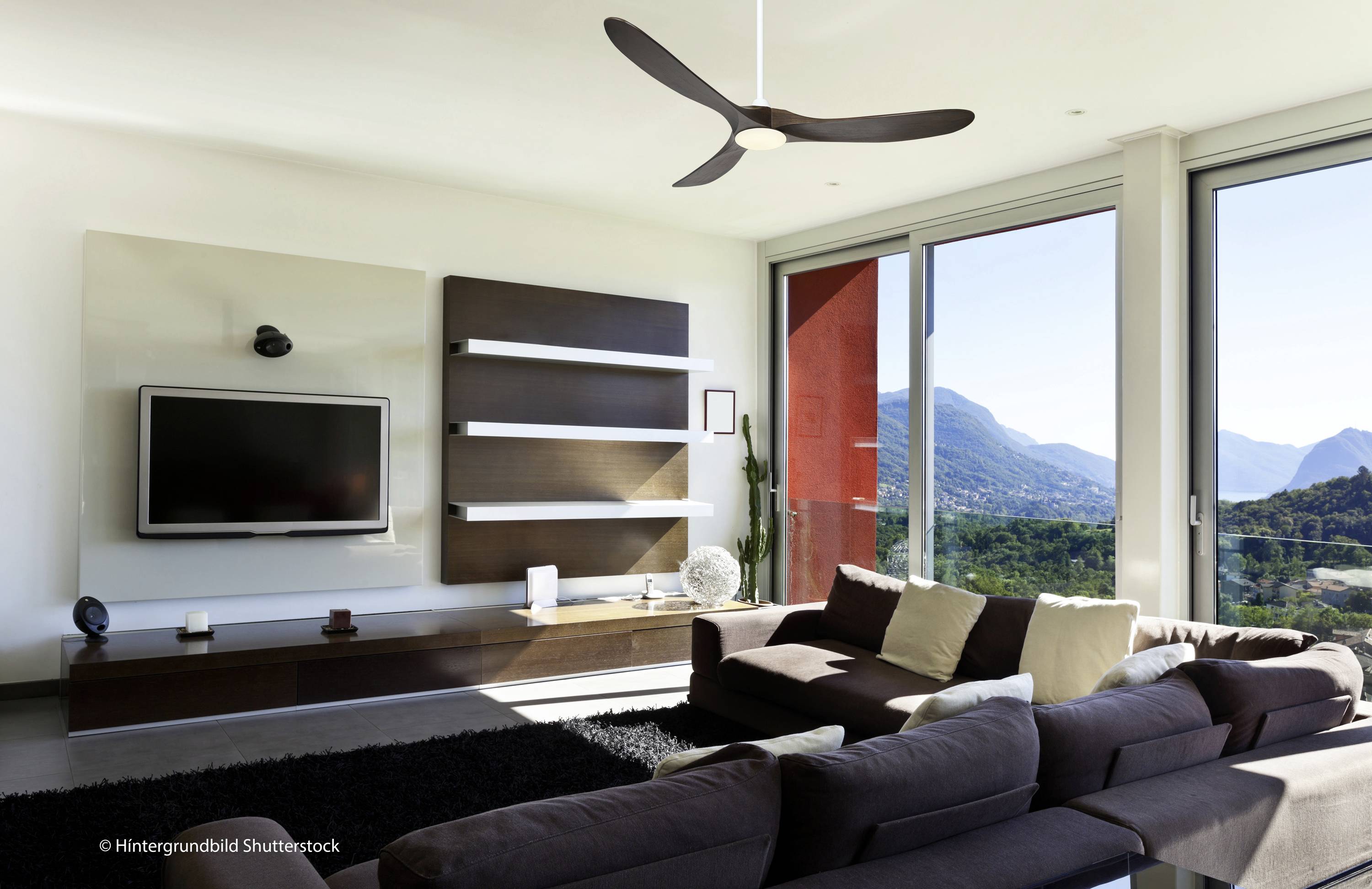 Modernes Wohnzimmer mit braunem Sofa, großem Fernseher, Regal und großem Fenster mit Aussicht auf Berge und Natur.