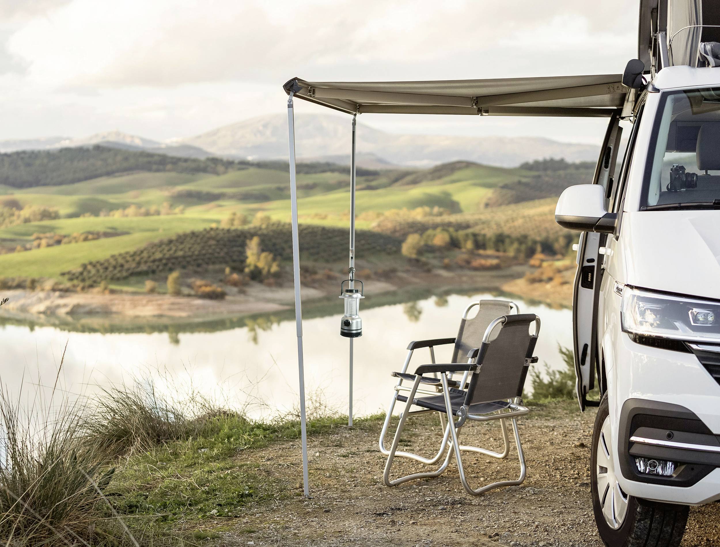 Wohnmobil mit Markise an einem Seeufer. Zwei Klappstühle stehen unter der Markise, im Hintergrund sanfte Hügel und ein ruhiger See.