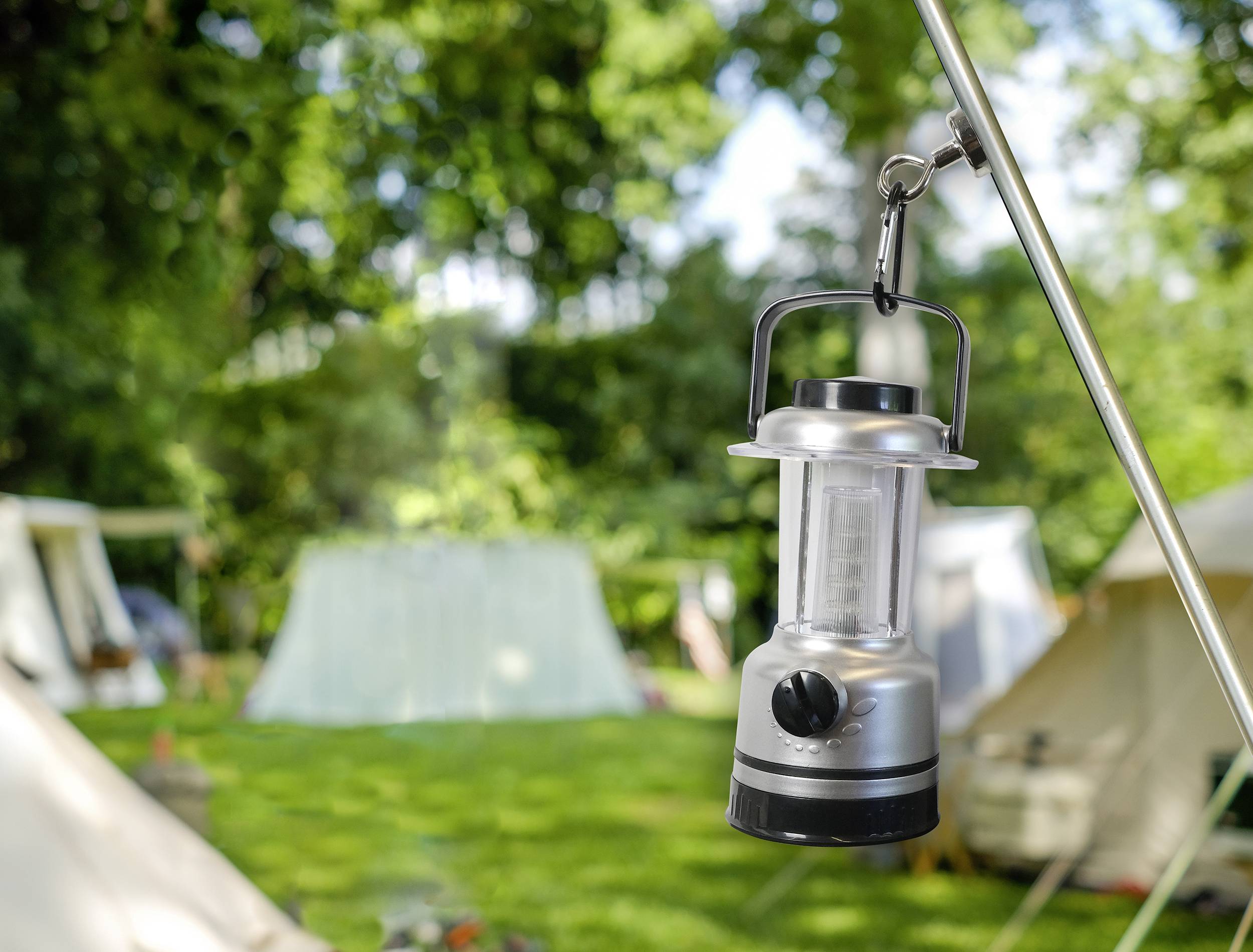 Eine silberne Laterne hängt an einer Zeltstange im Grünen. Im Hintergrund sind unscharf mehrere Zelte zu erkennen.