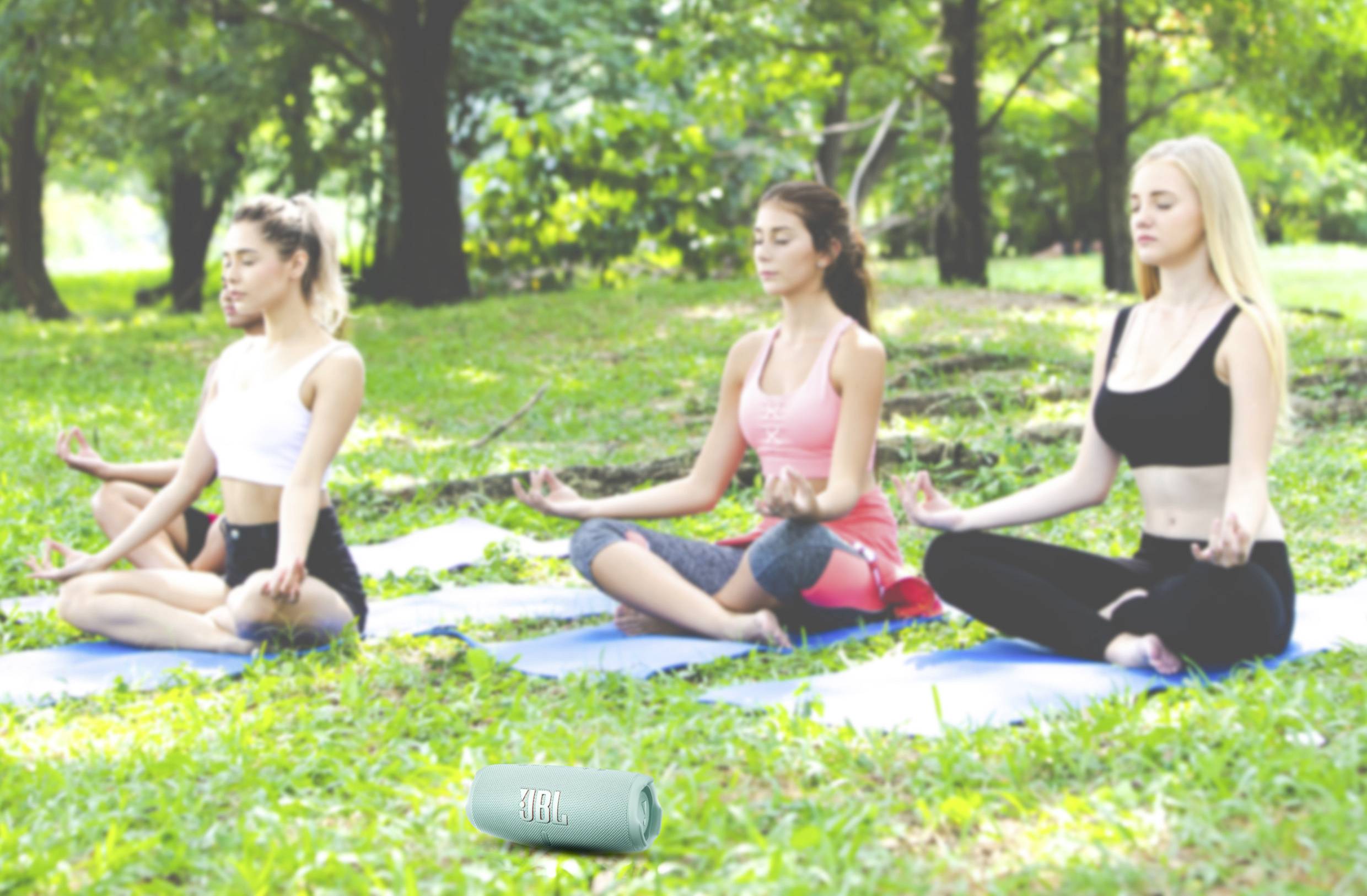 Drei Frauen sitzen im Freien auf Yogamatten und meditieren im Schneidersitz in einem parkähnlichen Umfeld.