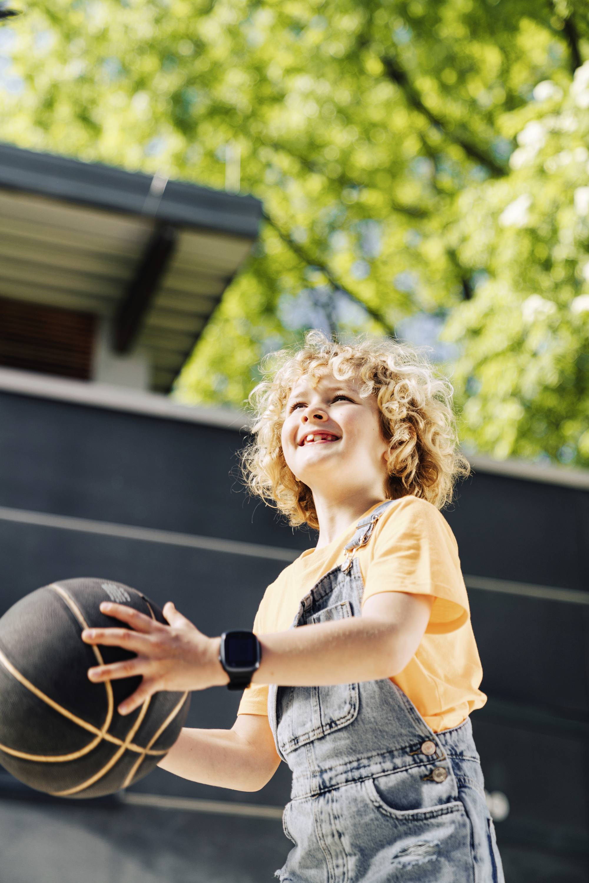 Ein fröhliches Kind mit lockigem Haar hält einen Basketball in einem sonnigen Park, umgeben von grünen Bäumen.