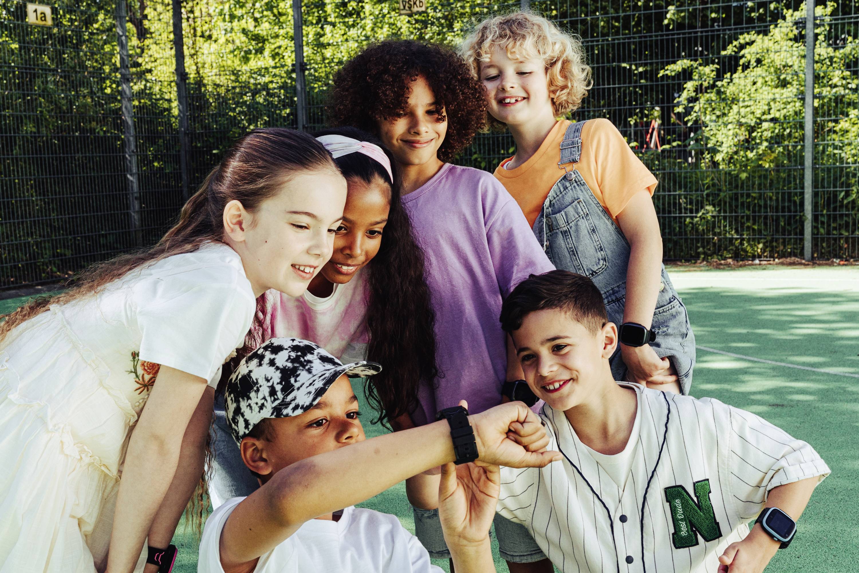 Eine Gruppe von sechs lächelnden Kindern steht eng beisammen auf einem Sportplatz. Ein Kind zeigt etwas auf einem Smartwatch-Display.