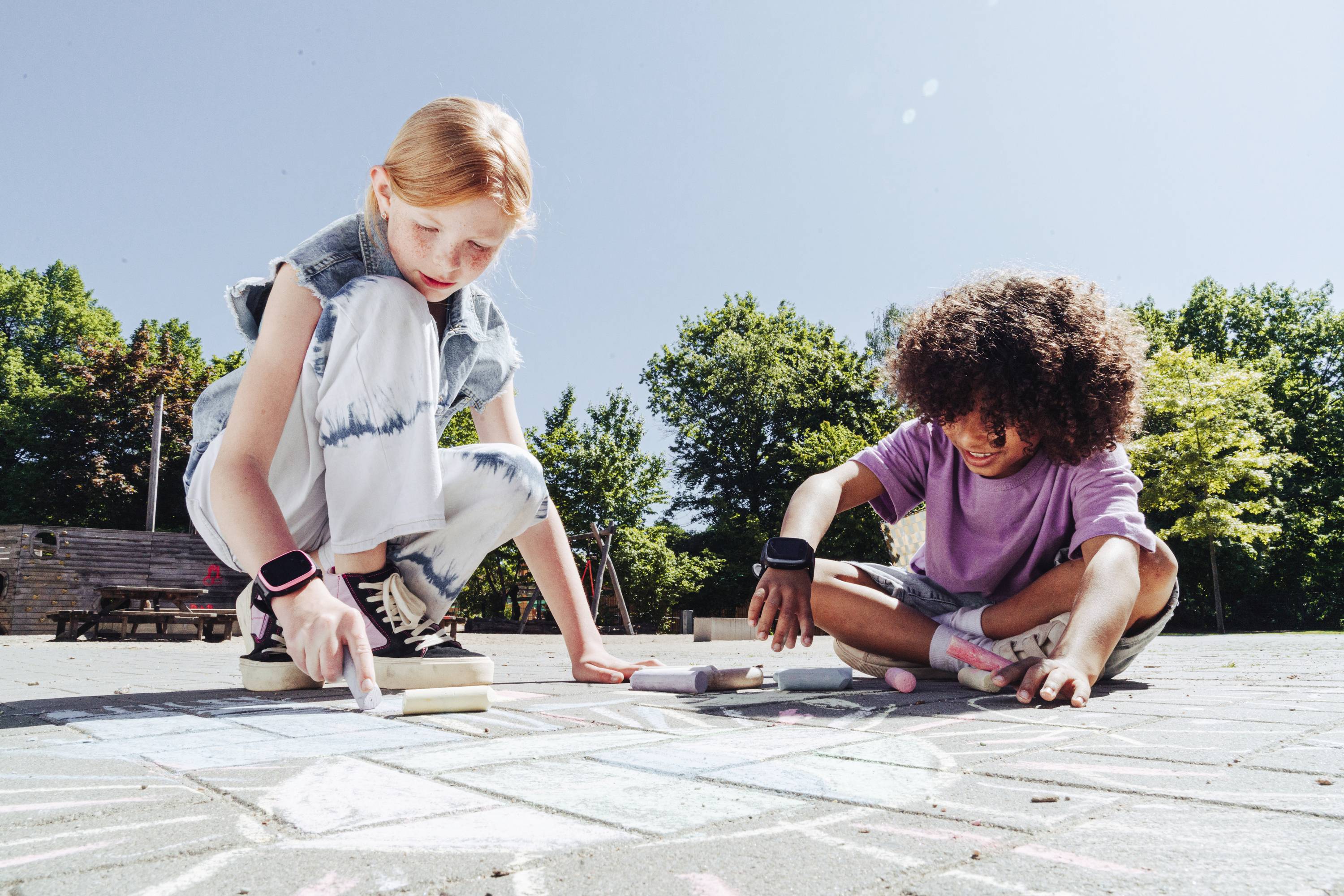 Zwei Kinder malen mit Kreide auf dem Boden eines sonnigen Spielplatzes. Beide tragen lässige Kleidung und wirken konzentriert.