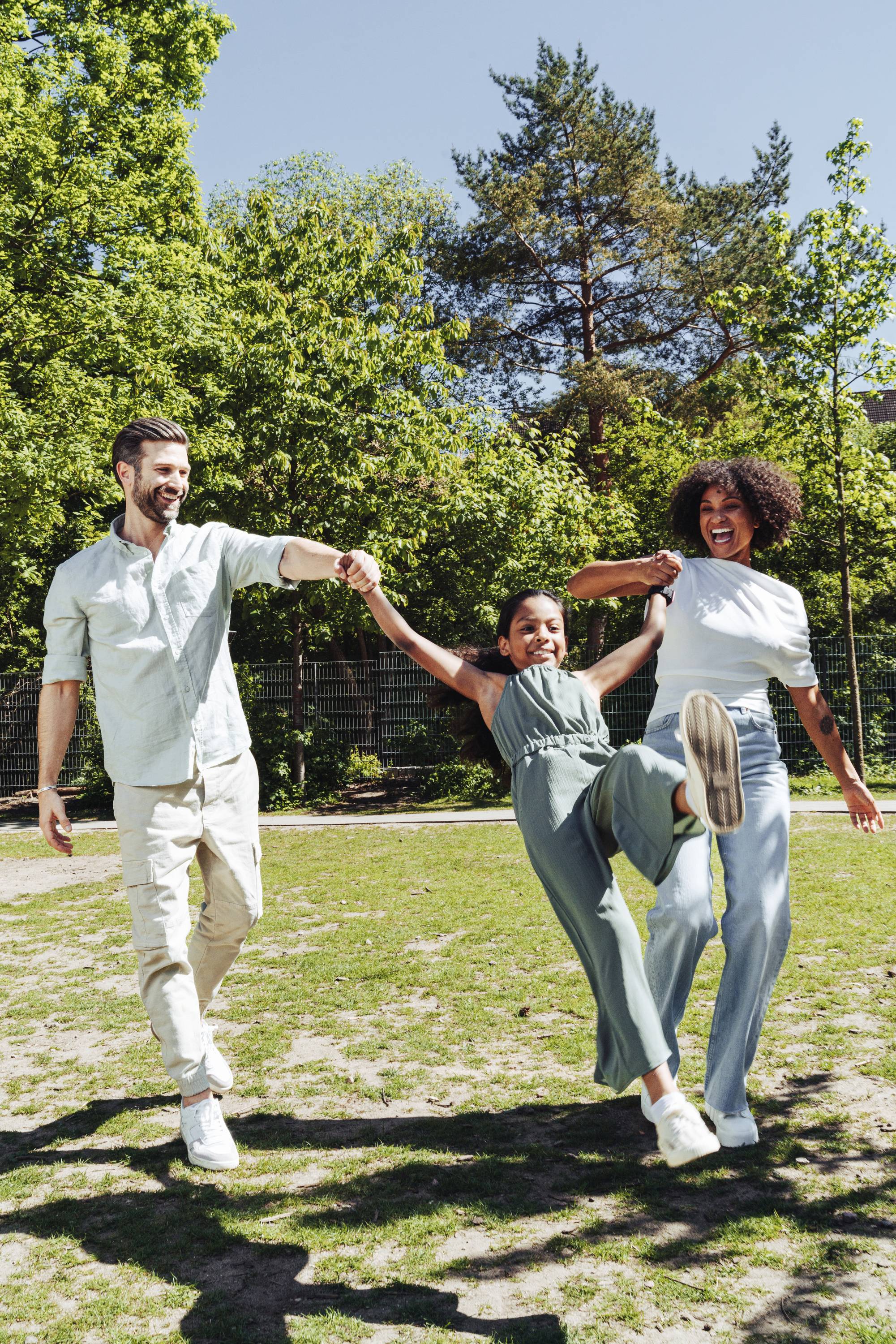 Eine lachende Familie schwingt ein Kind fröhlich in der Luft in einem sonnigen Park. Bäume und ein Zaun sind im Hintergrund sichtbar.