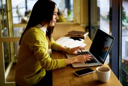 Frau sitzt an einem Laptop in einem Café, konzentriert arbeitend, mit Büchern, Handy, und einer Tasse Kaffee auf dem Tisch.
