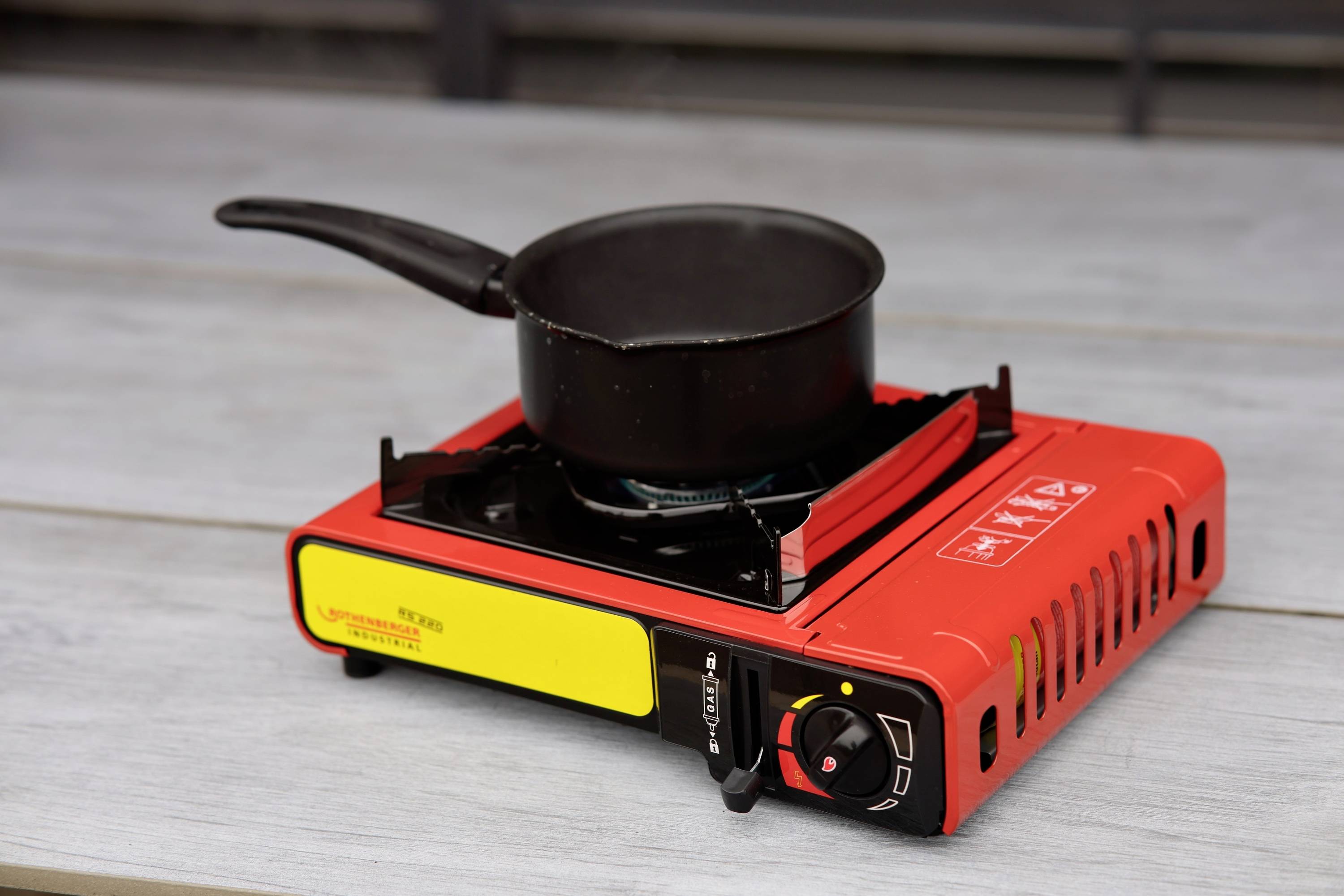 Ein kleiner roter Campingkocher mit schwarzem Topf steht auf einem Tisch. Der Kocher hat eine gelbe Beschriftung und Bedienknöpfe.