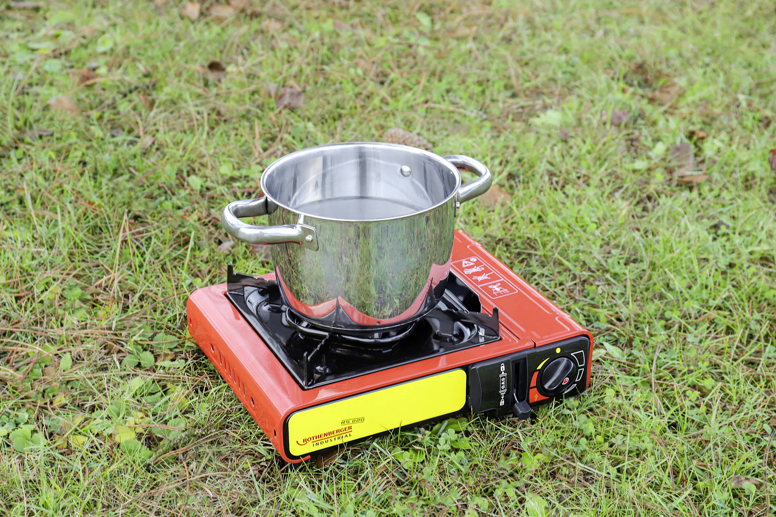 Ein Topf mit Wasser steht auf einem tragbaren roten Campingkocher im Gras.