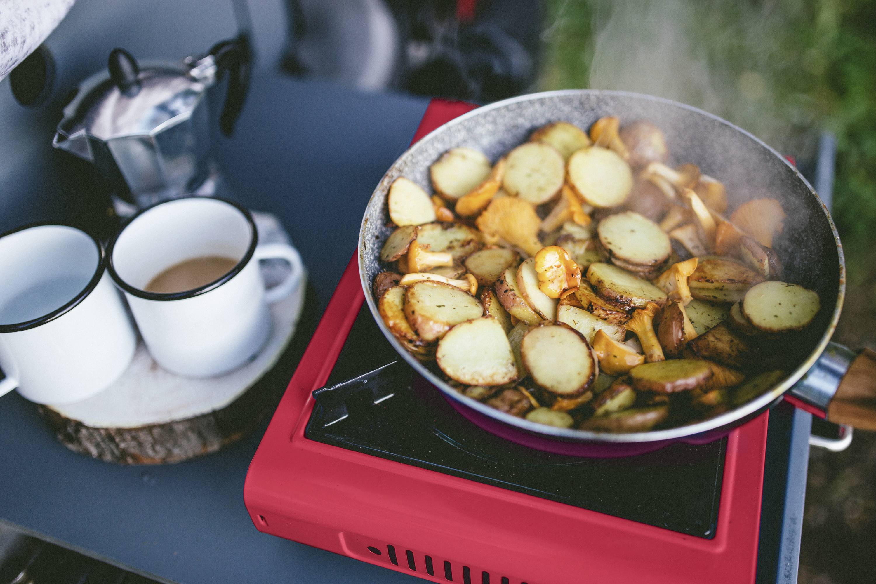 Kartoffelscheiben und Zwiebeln braten in einer Pfanne auf rotem Campingkocher. Daneben stehen zwei Emailtassen mit heißem Getränk.