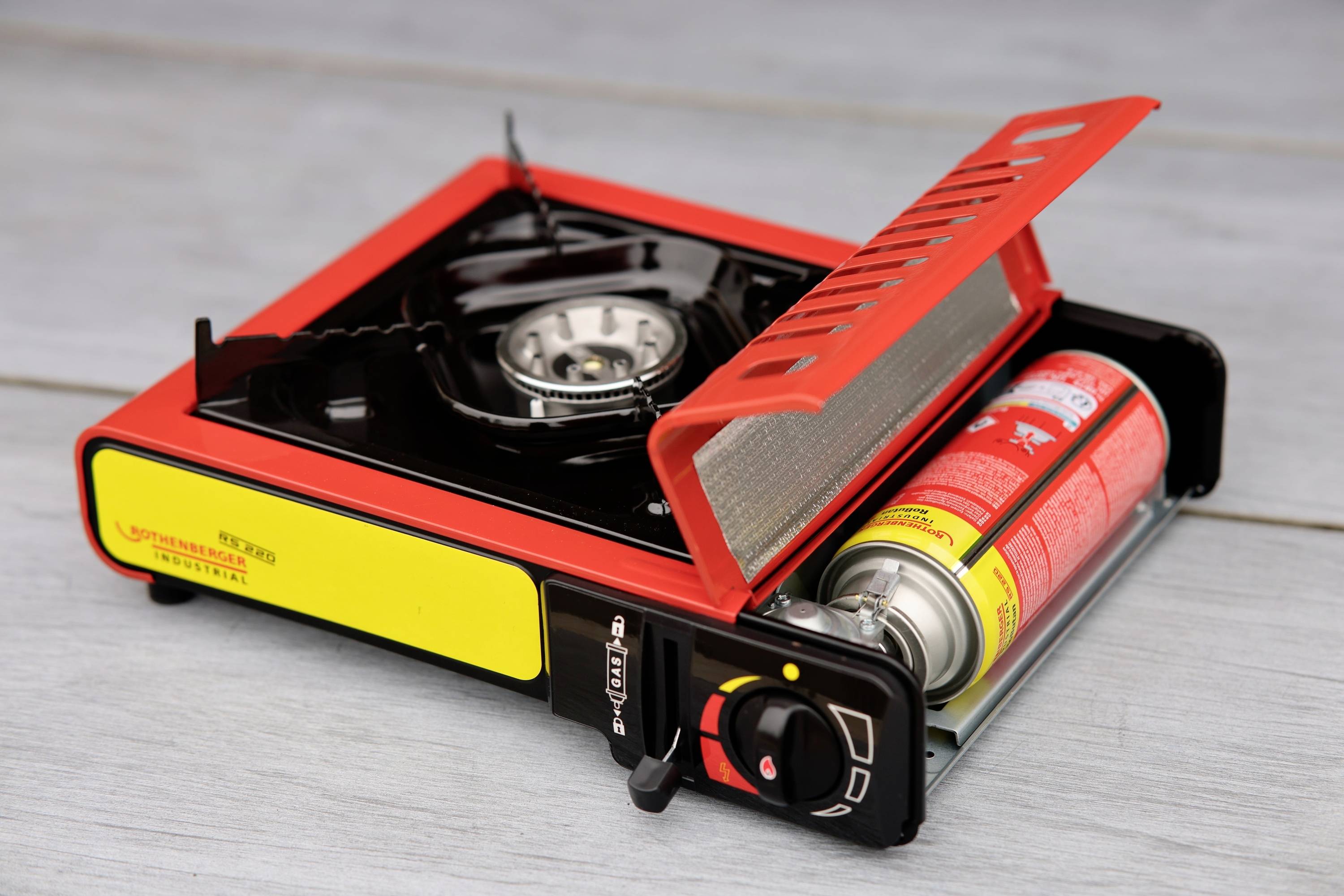 Ein tragbarer, rot-gelber Camping-Gaskocher mit eingesetzter Gaskartusche auf einem Holztisch. Der Kocher hat einen Flammenregler.