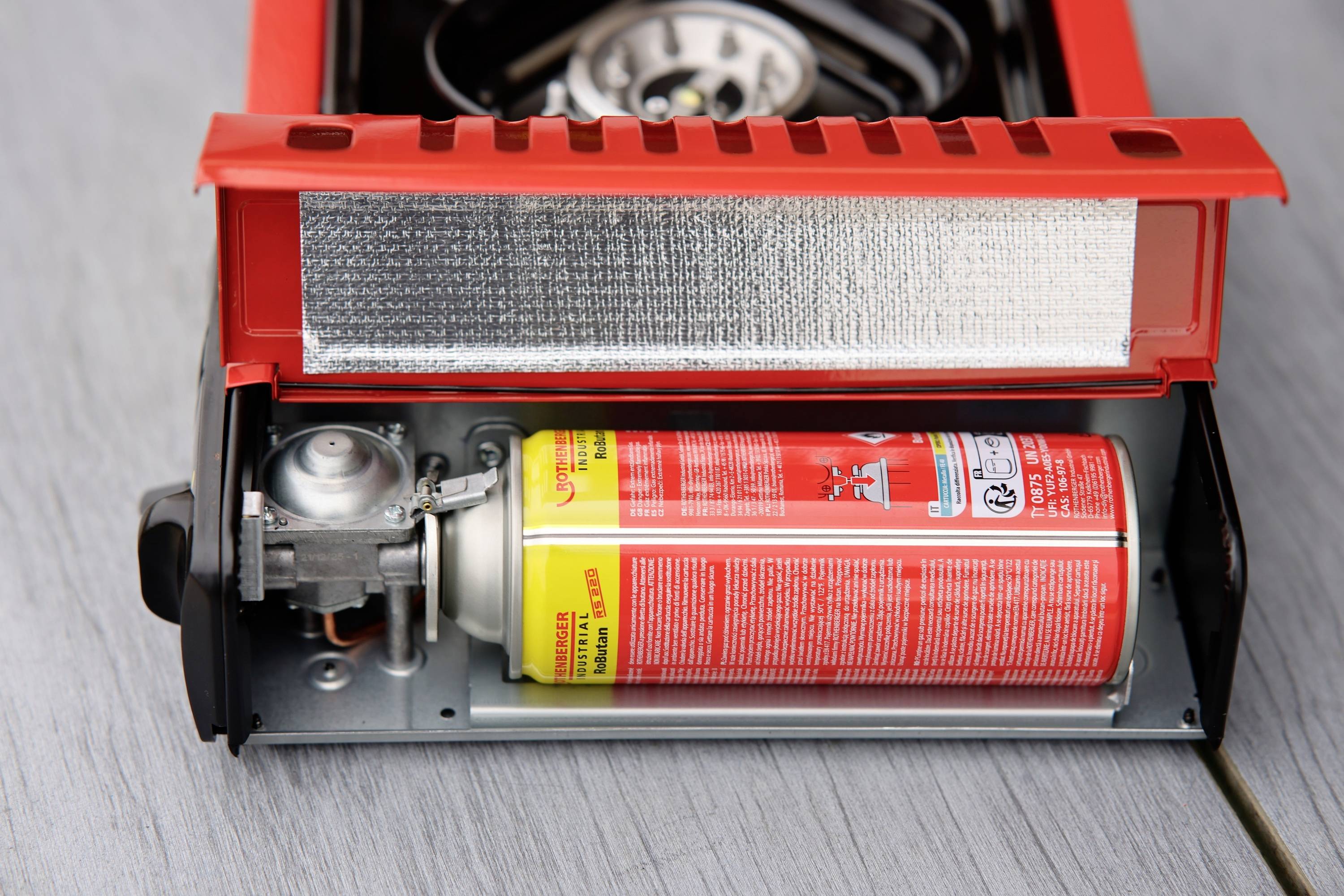 Ein roter tragbarer Gaskocher mit geöffneter Abdeckung zeigt eine eingelegte Butangas-Kartusche. Ideal für Campingausflüge.