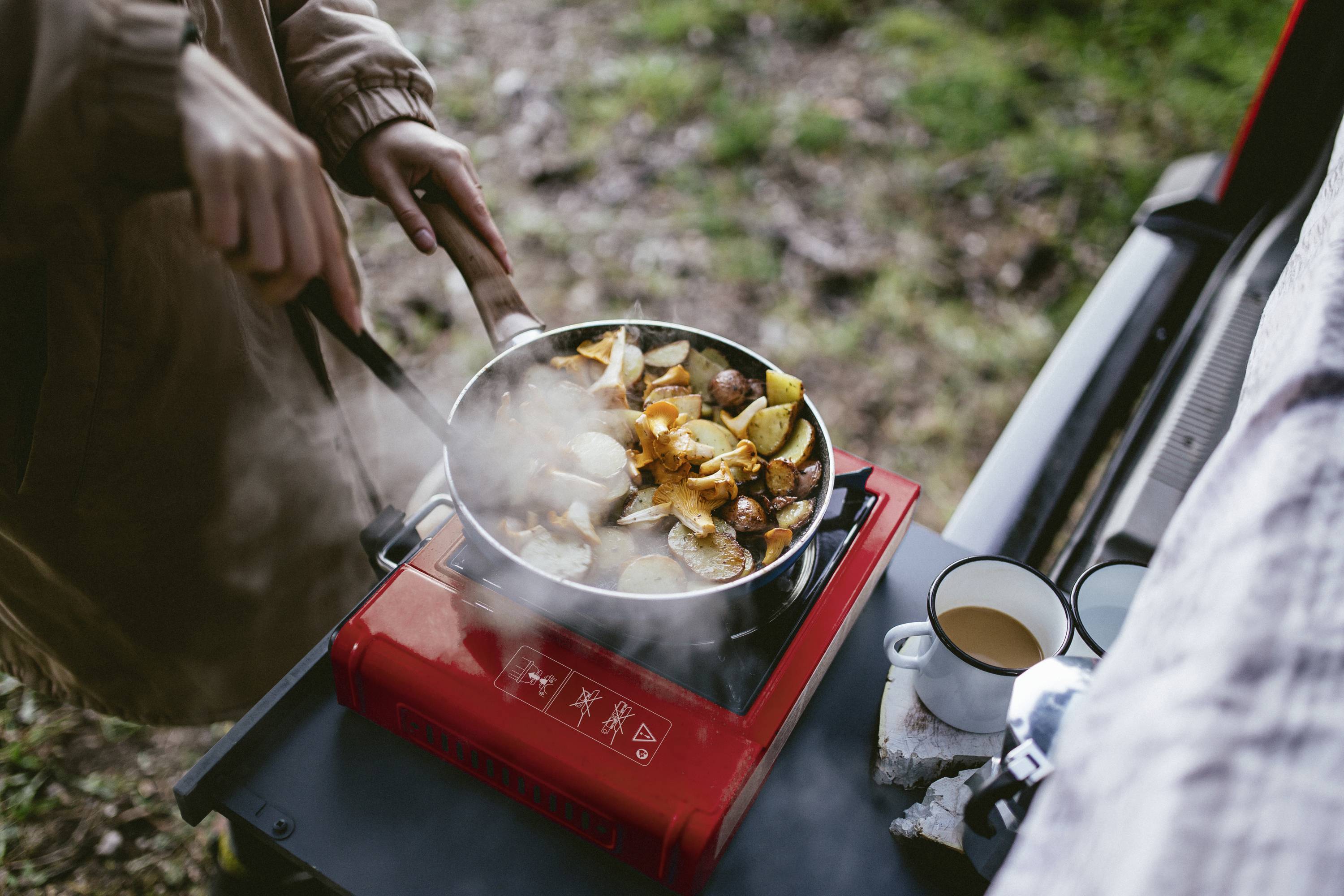 Eine Person kocht im Freien auf einem tragbaren Gaskocher eine Pfanne Gemüse und Würstchen. Daneben stehen zwei Tassen.
