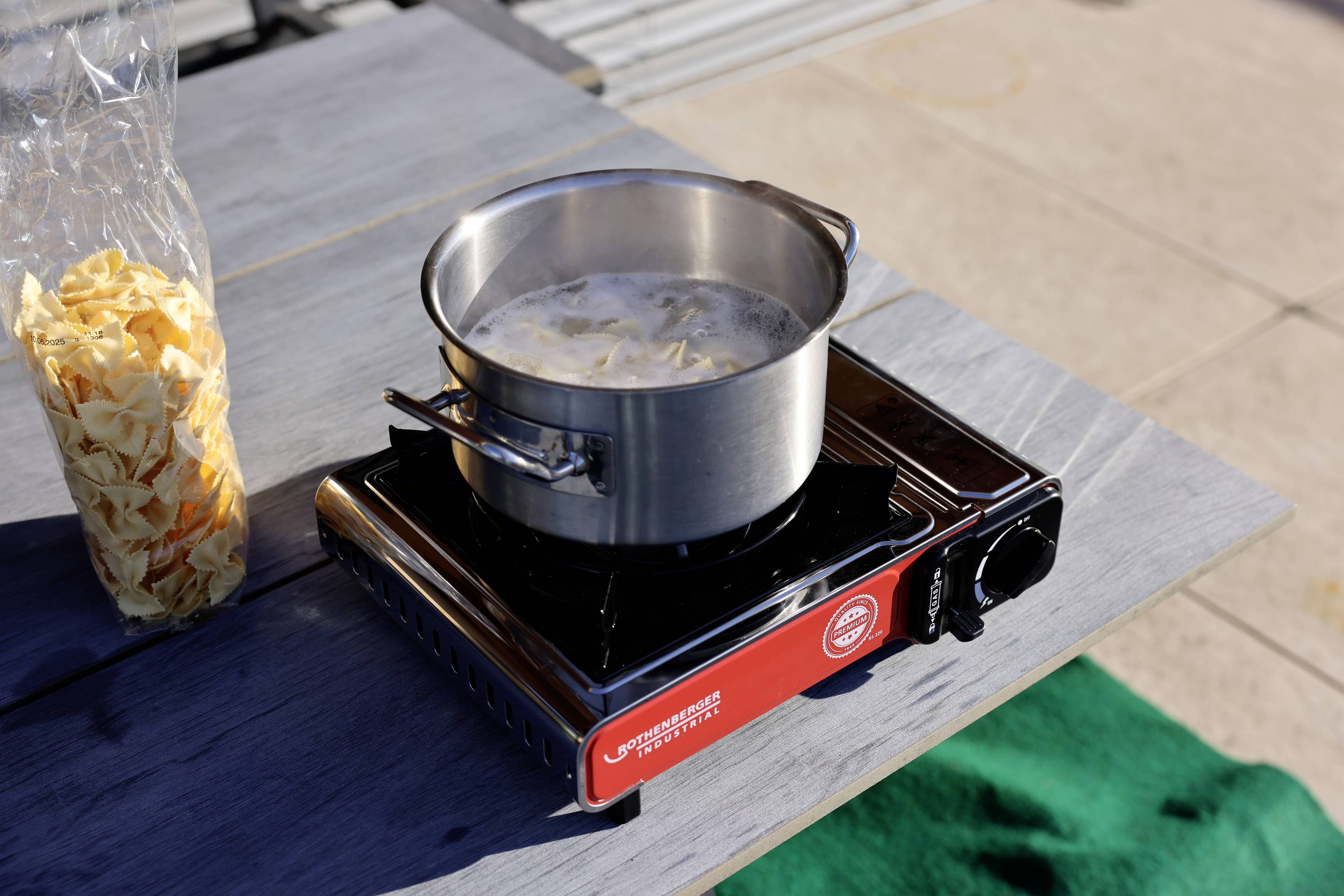 Ein Topf mit kochendem Wasser und Nudeln steht auf einem tragbaren Gasherd im Freien. Daneben liegt eine geöffnete Nudelpackung.