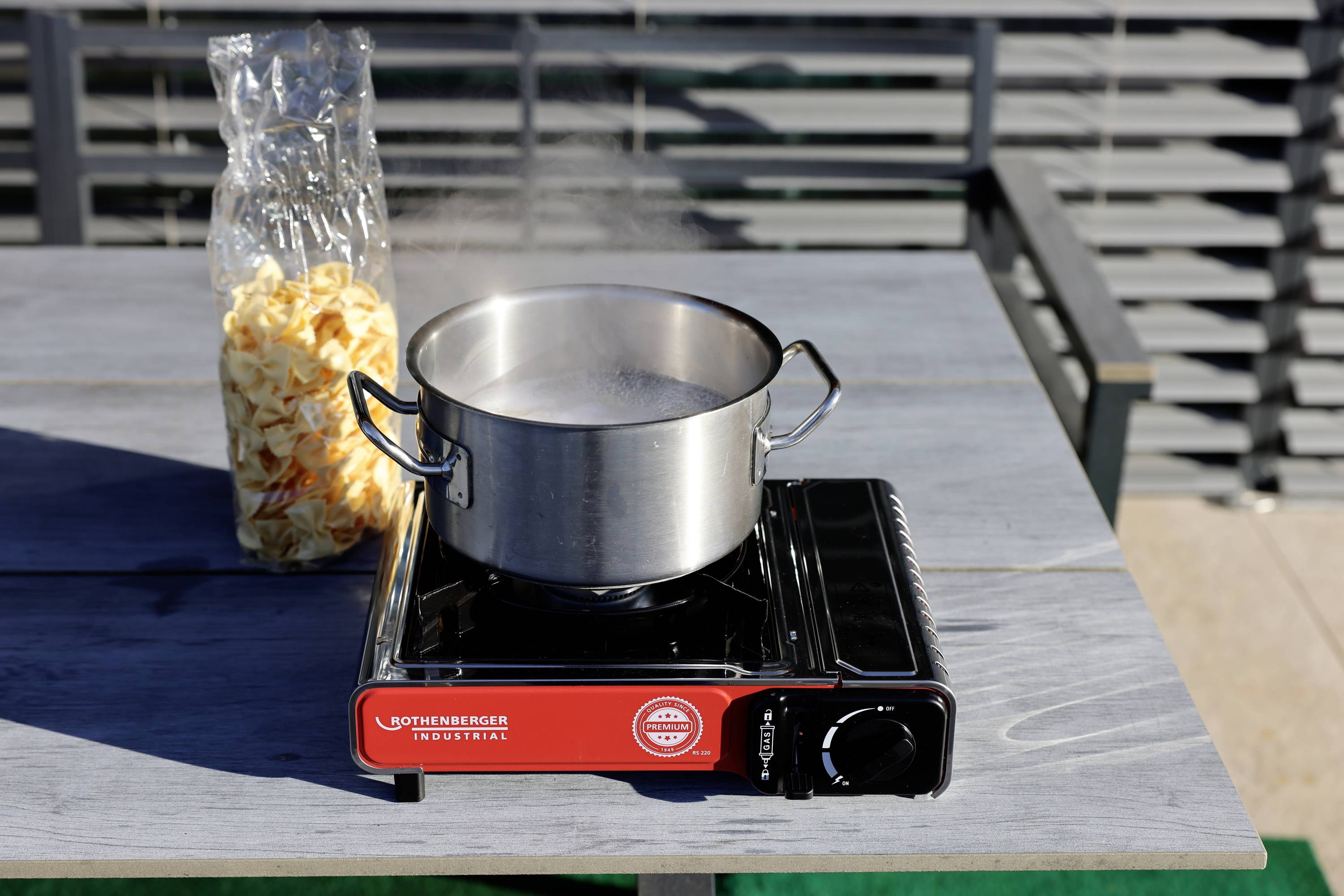 Ein Topf kocht auf einem tragbaren Gasherd im Freien. Daneben steht eine geöffnete Tüte mit getrockneten Nudeln auf dem Tisch.