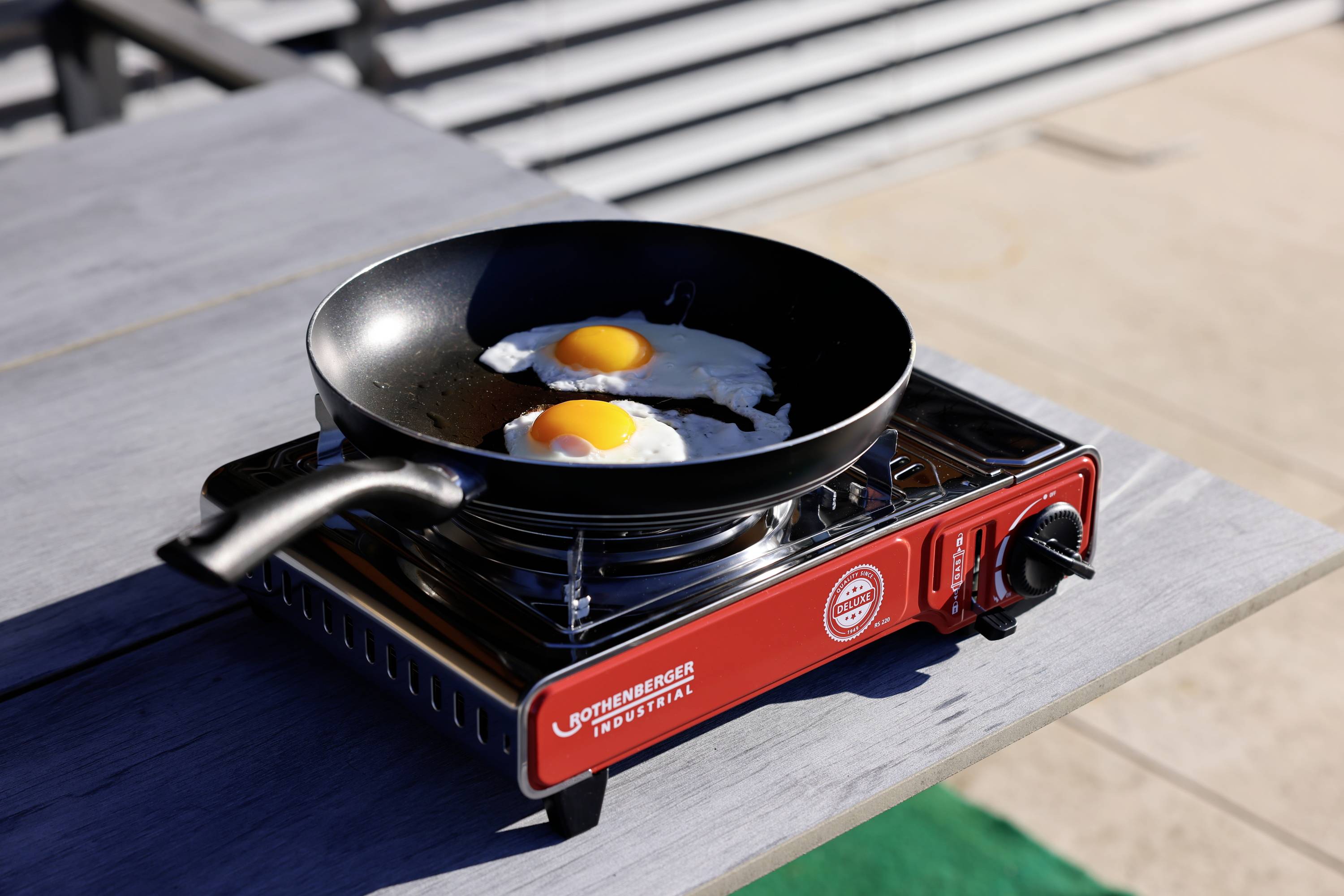 Eine Bratpfanne mit zwei Spiegeleiern steht auf einem roten tragbaren Gasherd auf einem Tisch im Freien.