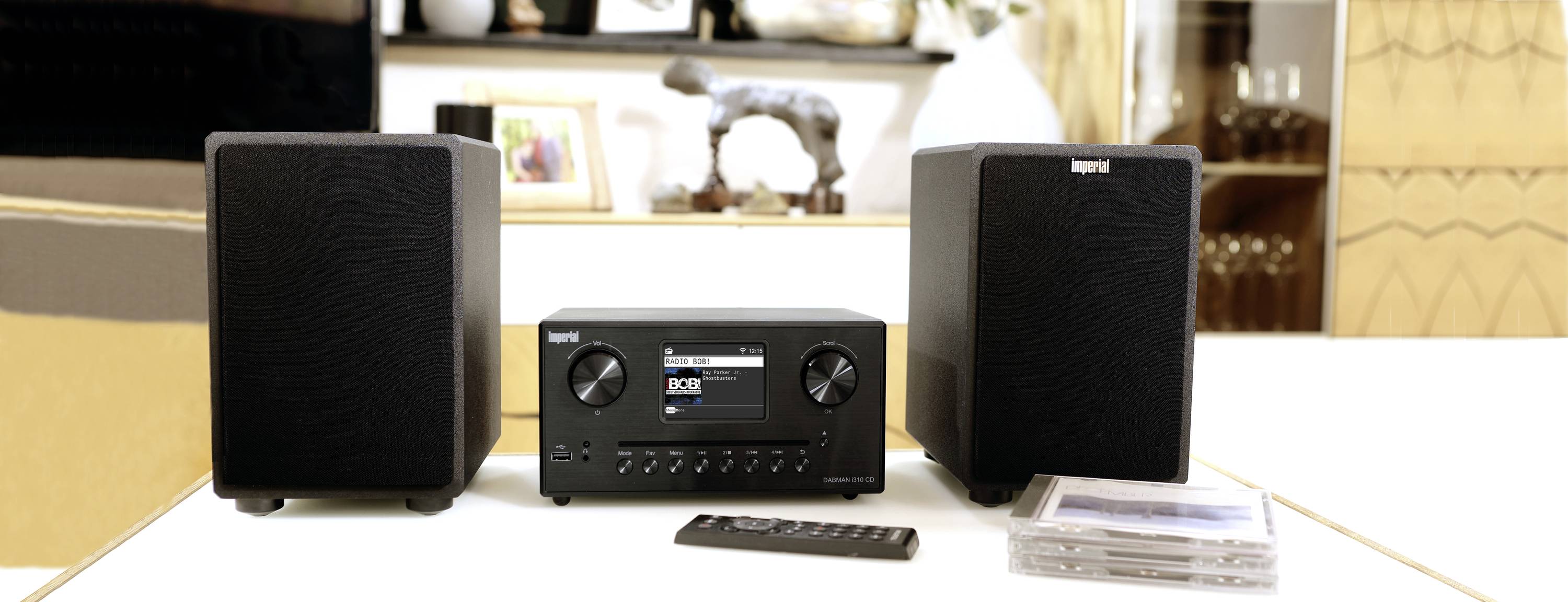 Ein schwarzes Stereo-Audiosystem mit zwei Lautsprechern, einem Hauptgerät mit Display und Drehknöpfen; eine Fernbedienung und CDs daneben.