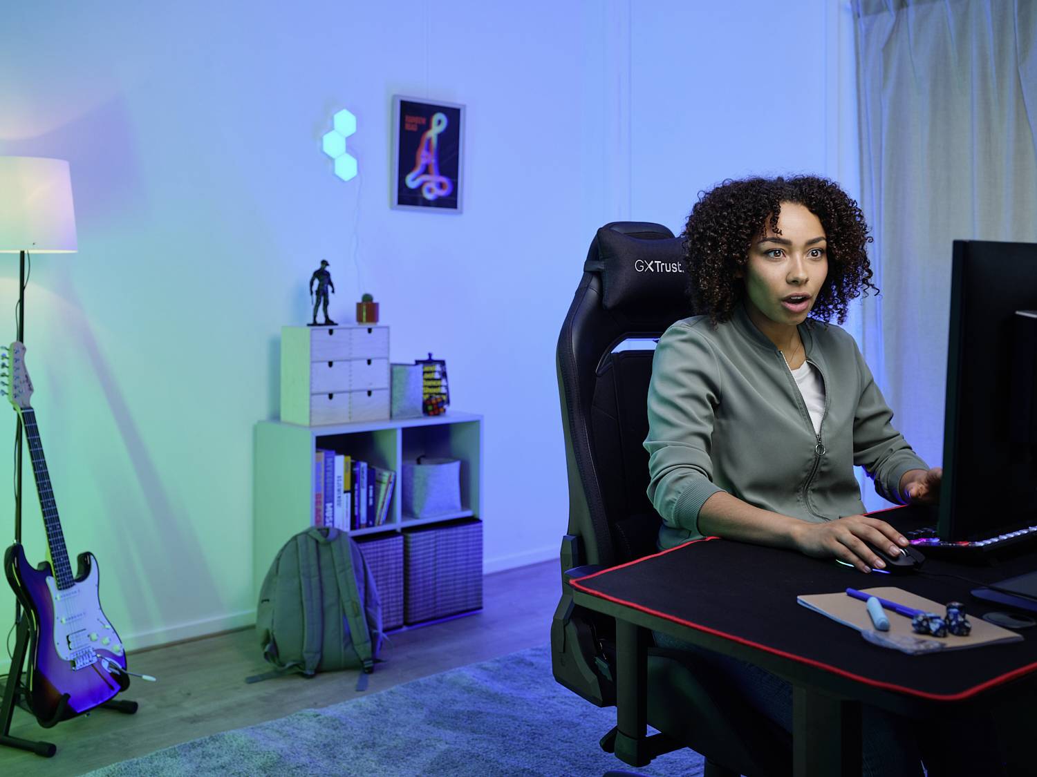 Eine Person sitzt an einem Schreibtisch vor einem Computerbildschirm in einem modern eingerichteten Raum mit Gitarre und Regal im Hintergrund.