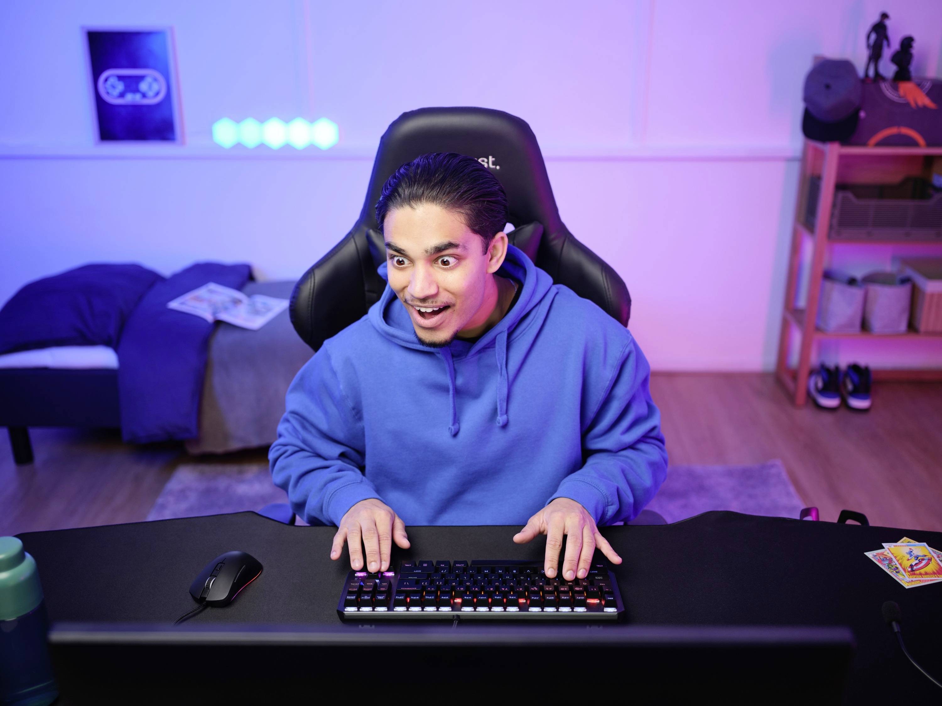Eine Person in einem blauen Hoodie sitzt an einem Schreibtisch mit Tastatur und zeigt ein freudiges Gesicht, möglicherweise beim Spielen am Computer.
