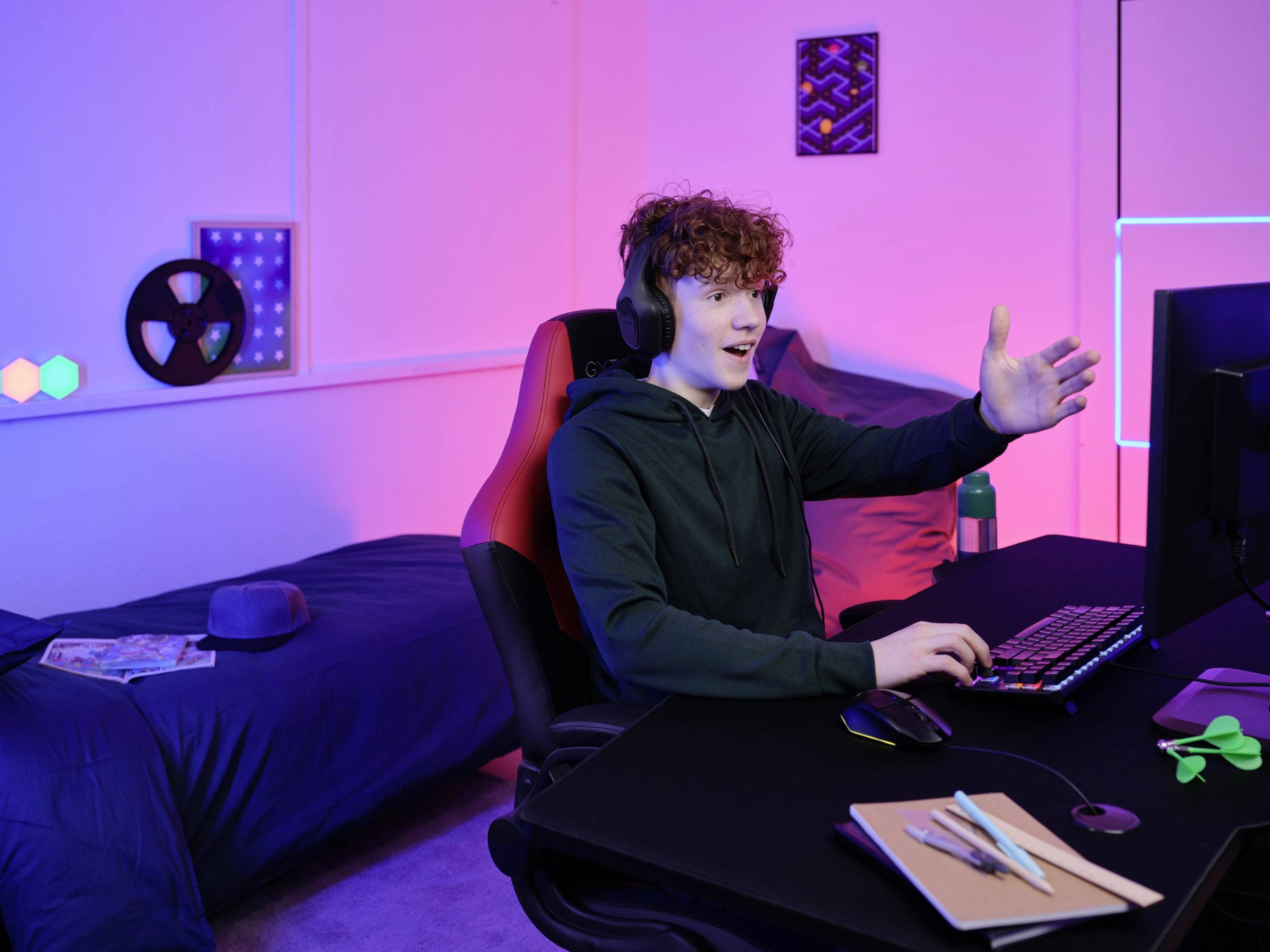 Ein junger Mann sitzt an einem Schreibtisch und spielt auf einem Computer, trägt Kopfhörer. Im Hintergrund ist ein Bett zu sehen.