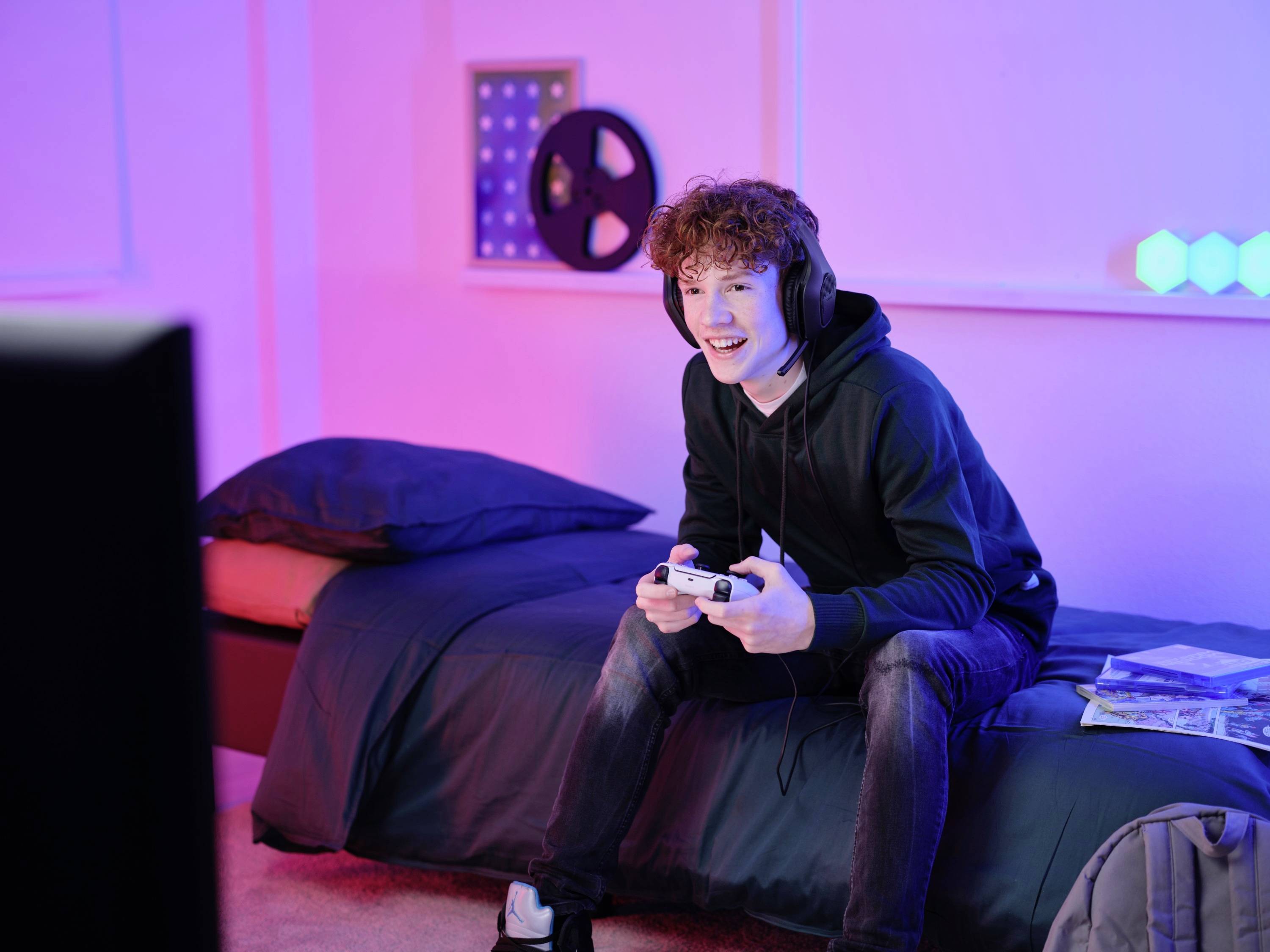 Ein junger Mann sitzt lächelnd auf einem Bett und spielt Videospiele. Er trägt Kopfhörer und hält einen Controller in der Hand.