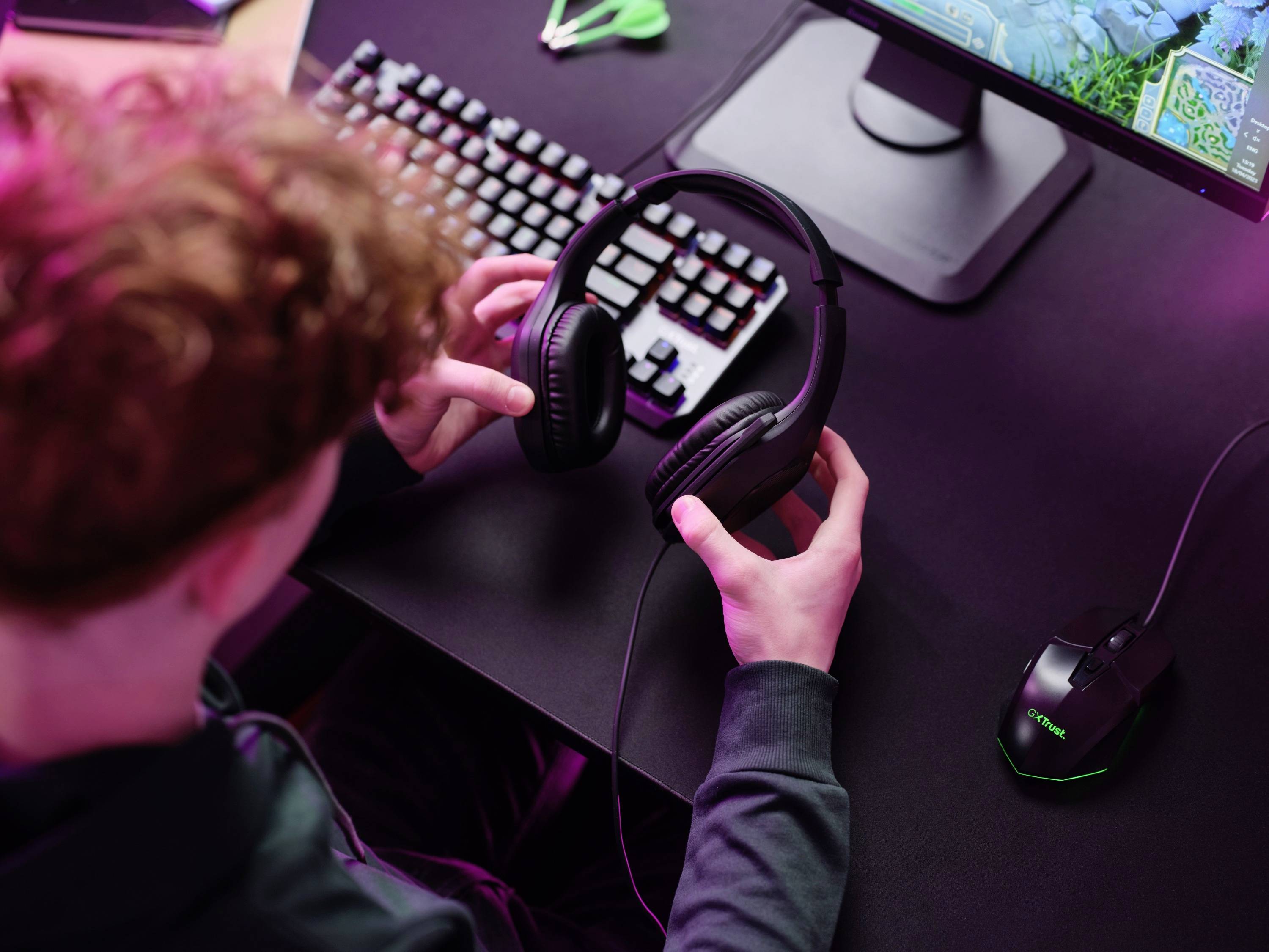 Eine Person sitzt an einem Tisch mit einem Computer, hält ein Headset in den Händen. Eine bunte Tastatur und Maus sind sichtbar.