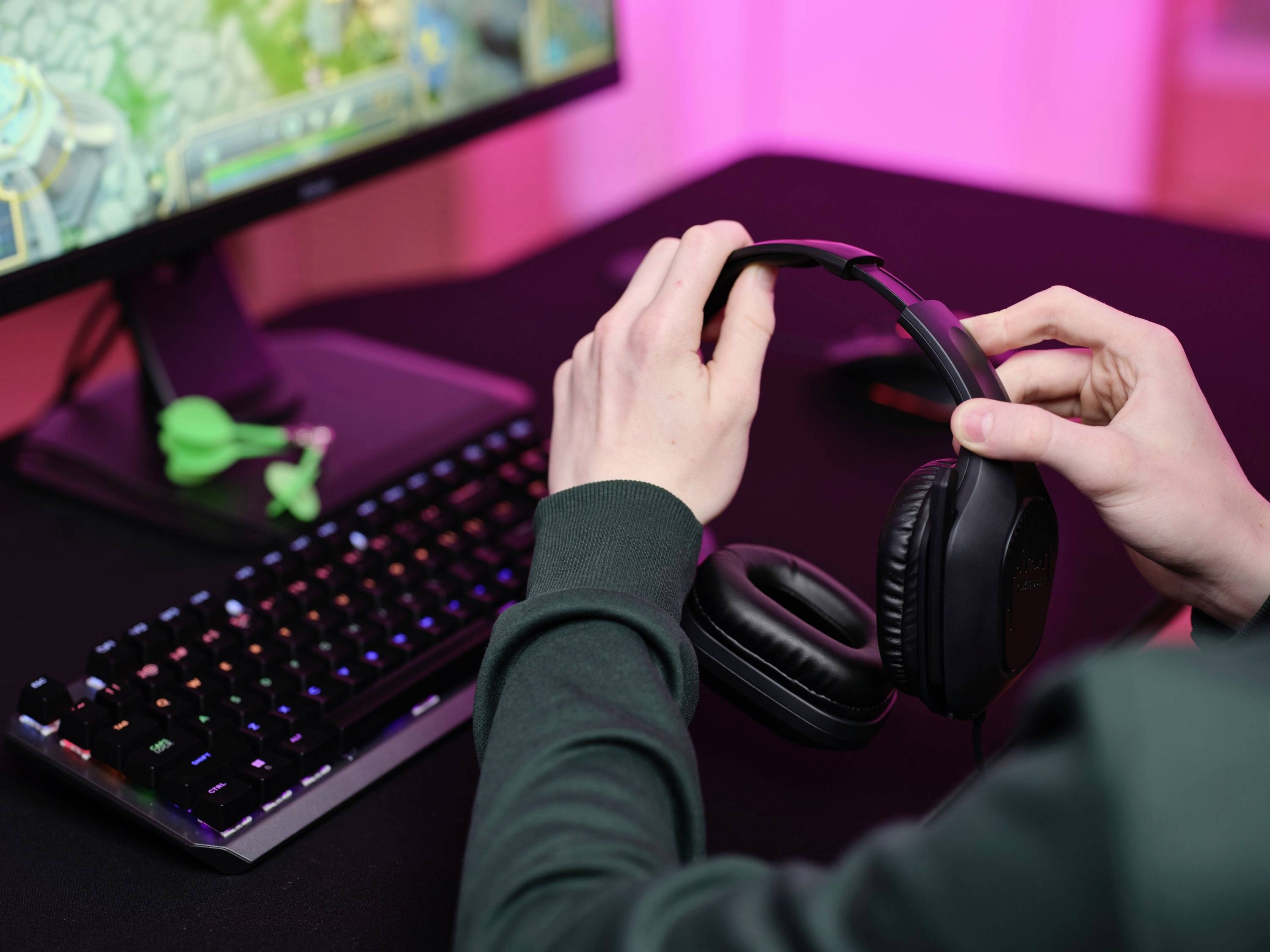 Person legt Kopfhörer ab, ein Computerbildschirm mit Videospiel erscheint im Hintergrund. Tastatur und Maus sind auf dem Tisch.