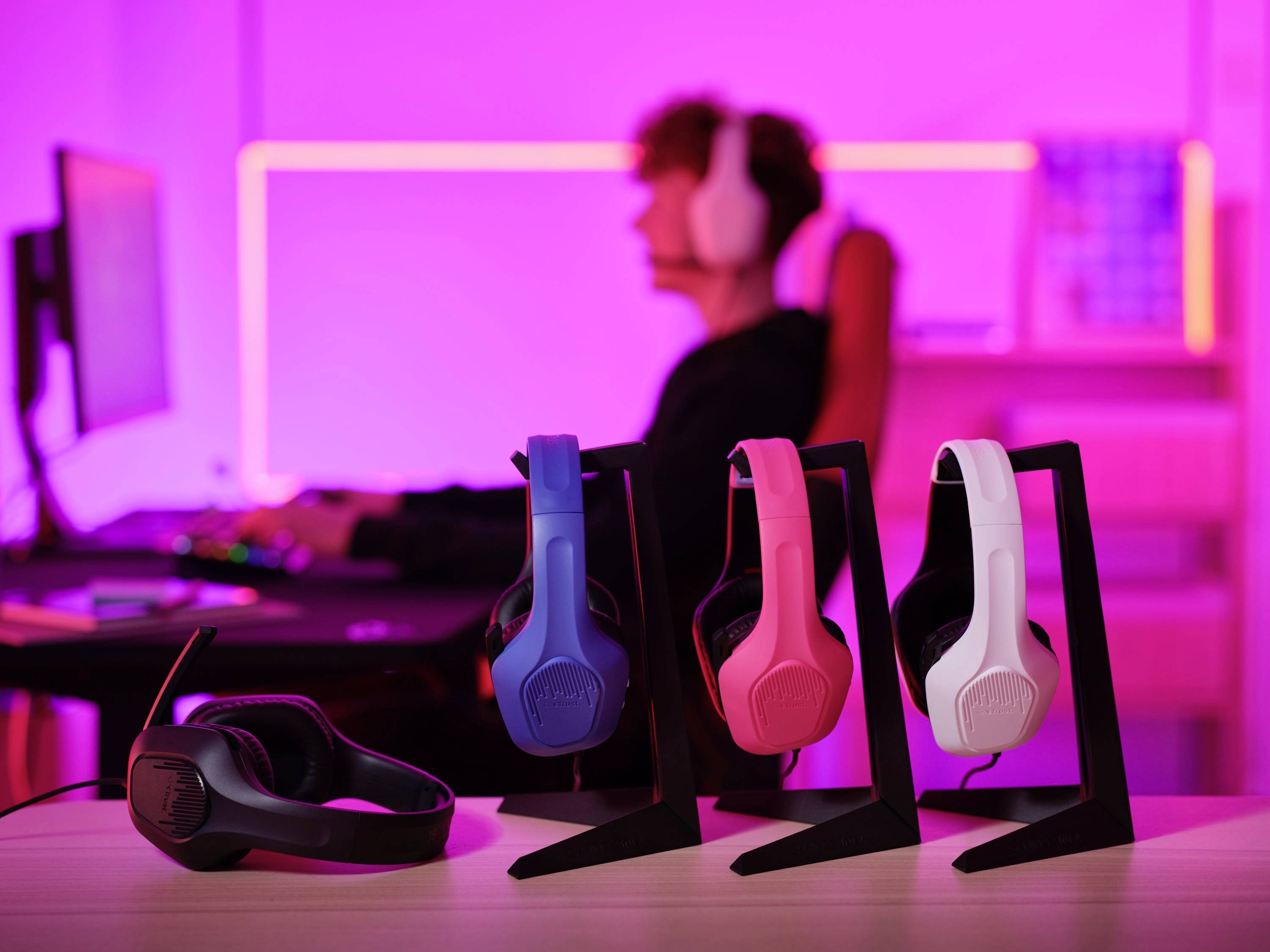 Vier bunte Kopfhörer auf Ständern vor einem unscharfen Hintergrund, auf dem eine Person vor einem Computer sitzt.